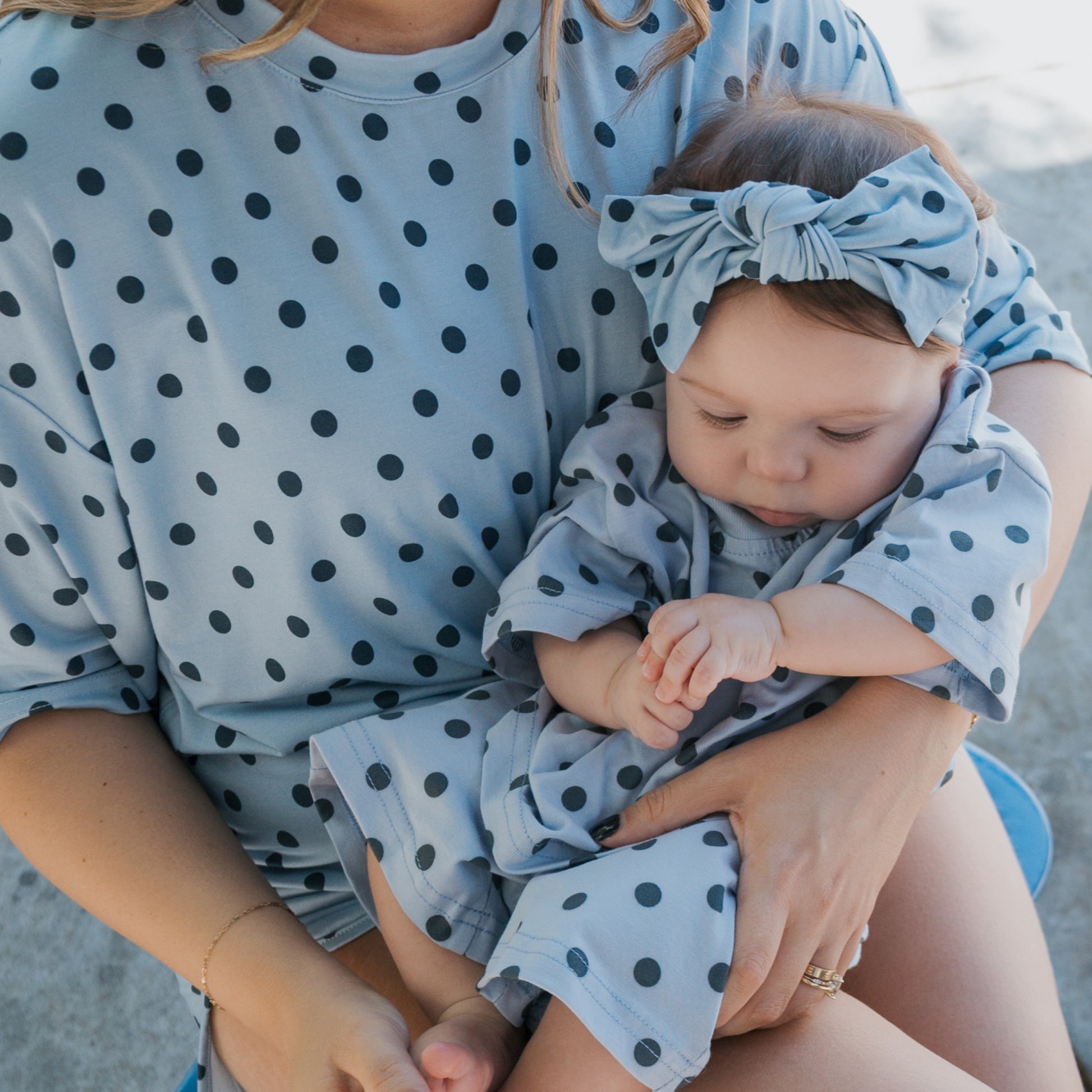 A woman holds a baby in her lap. Both are wearing matching light blue outfits with black polka dots. The baby, looking down at their hands, is sporting the Bamboo Head Wrap | Sully from forever french baby, complete with an adorable bow headband.