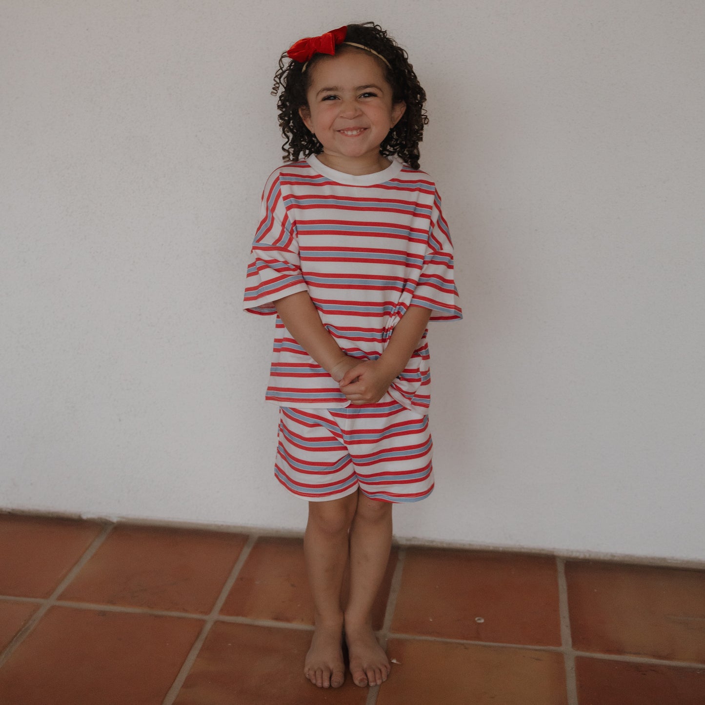 A young girl with curly hair and a red bow stands barefoot on a tiled floor, smiling. She wears the forever french baby Child Short Set | Heritage Stripe in red, white, and blue against a plain white wall.