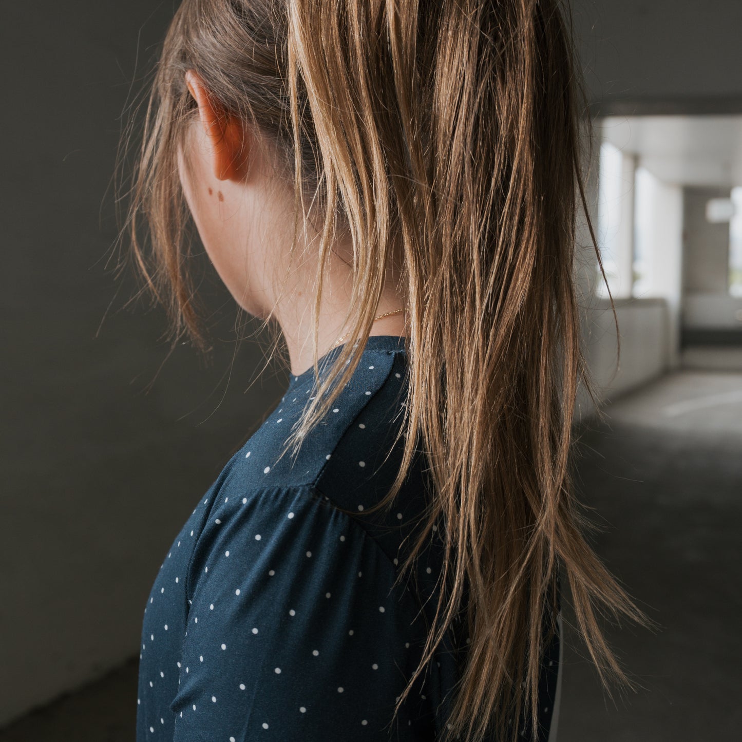 A child with long, light brown hair in a ponytail faces away indoors, wearing the forever french baby Child Short Sleeve Bamboo Lounge Set | Emery—a dark blue set with small white polka dots. The softly lit background is blurred.