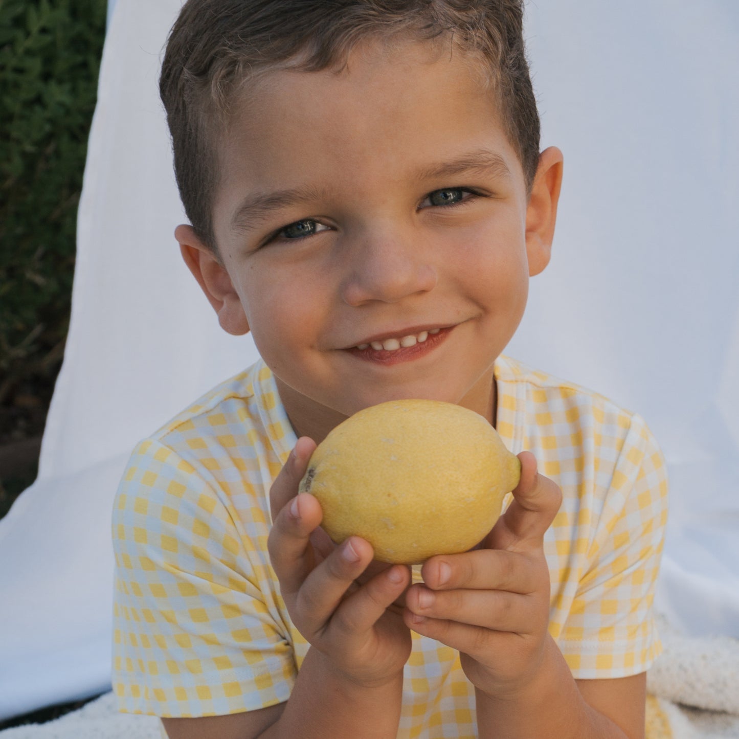 A young boy with short brown hair, wearing the forever french baby Bamboo Short Set in Citron, smiles and holds a large yellow lemon with both hands. A white cloth and greenery are visible in the background.