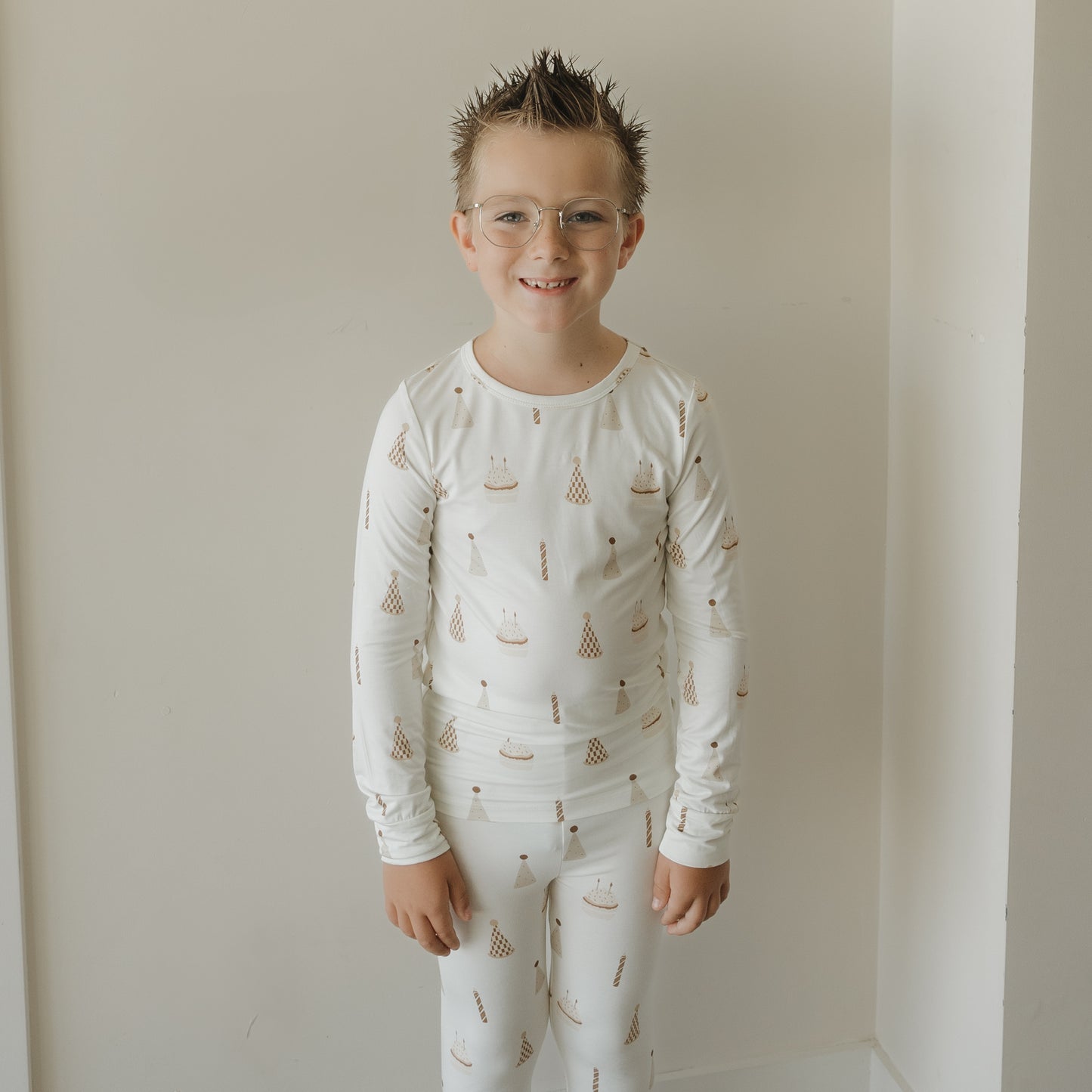 A young boy with spiky hair and glasses smiles while wearing the forever french baby Child Sleepy Time Set | Birthday Boy, featuring breathable fabric with small birthday cake and party hat patterns, standing against a plain white wall.