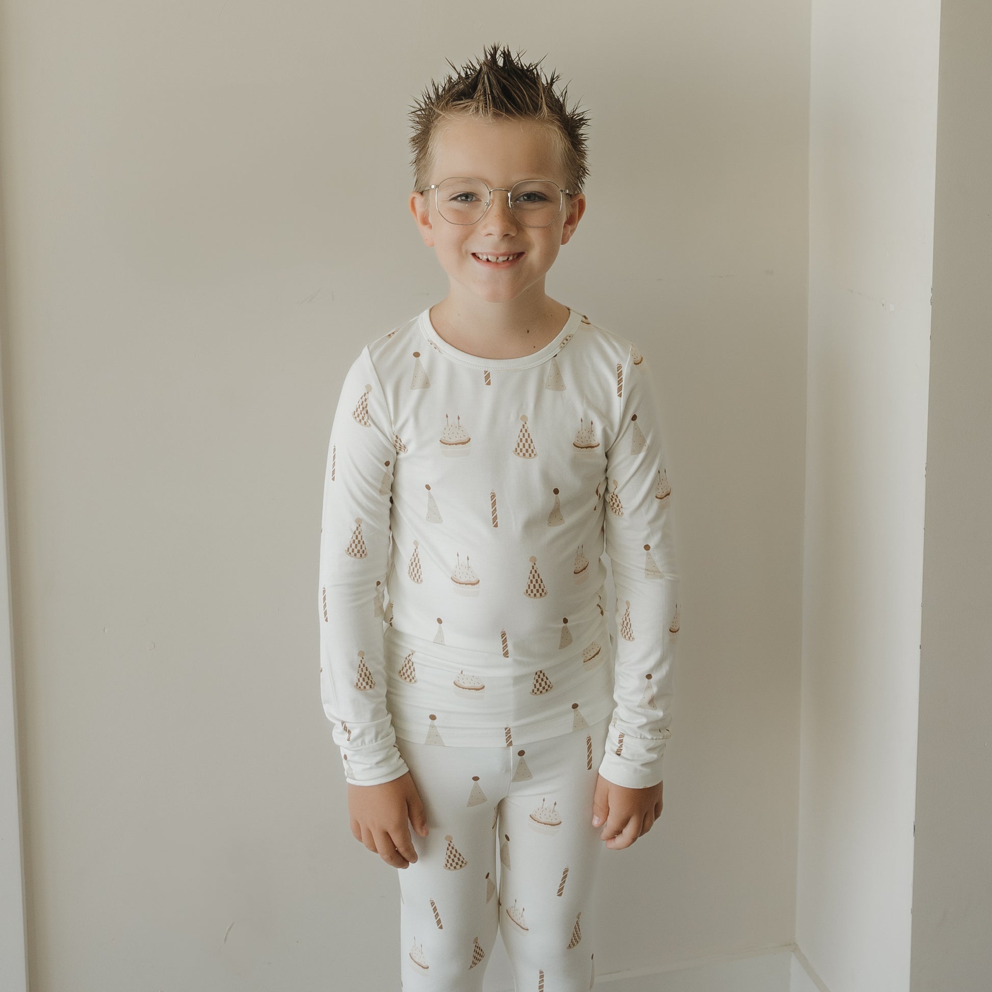 A young boy with spiky hair and glasses smiles while wearing the forever french baby Child Sleepy Time Set | Birthday Boy, featuring breathable fabric with small birthday cake and party hat patterns, standing against a plain white wall.