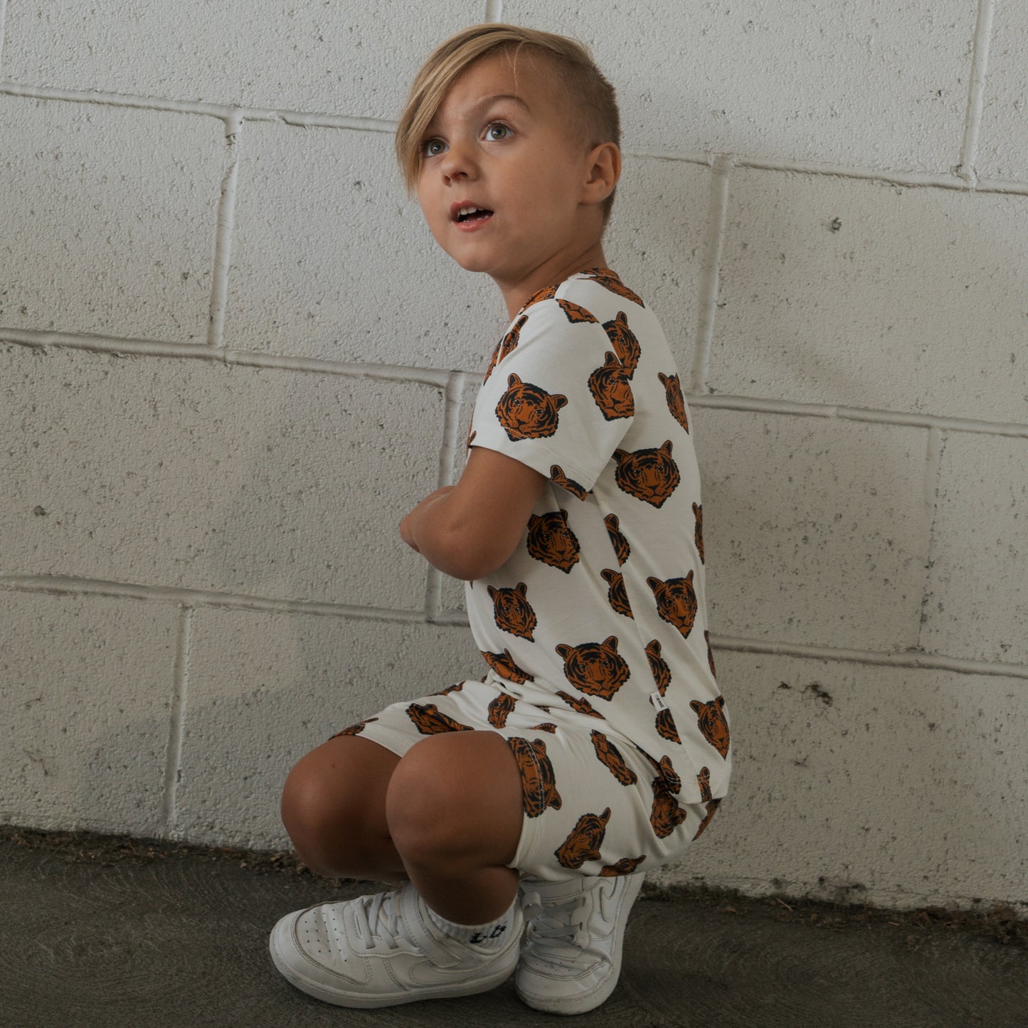 A young child with blonde hair squats by a white cinder block wall, wearing the forever french baby Bamboo Short Set | Tigers and white sneakers, looking slightly upward with an open-mouthed expression.