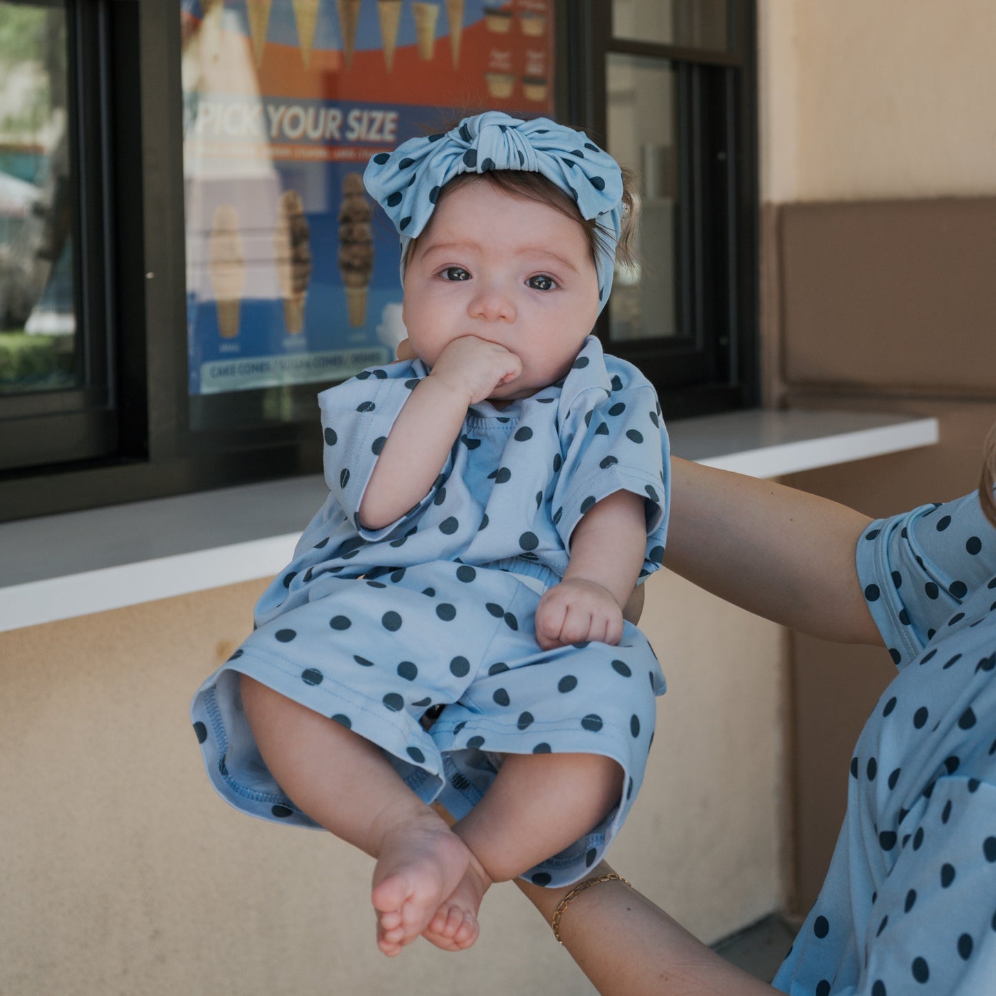 A baby wearing a blue polka-dotted outfit and the Bamboo Head Wrap | Sully from forever french baby is held up by an adult. The baby has a hand in their mouth, adding to the adorable moment. An ice cream poster is visible in the background, perfectly capturing this sweet forever french baby scene.