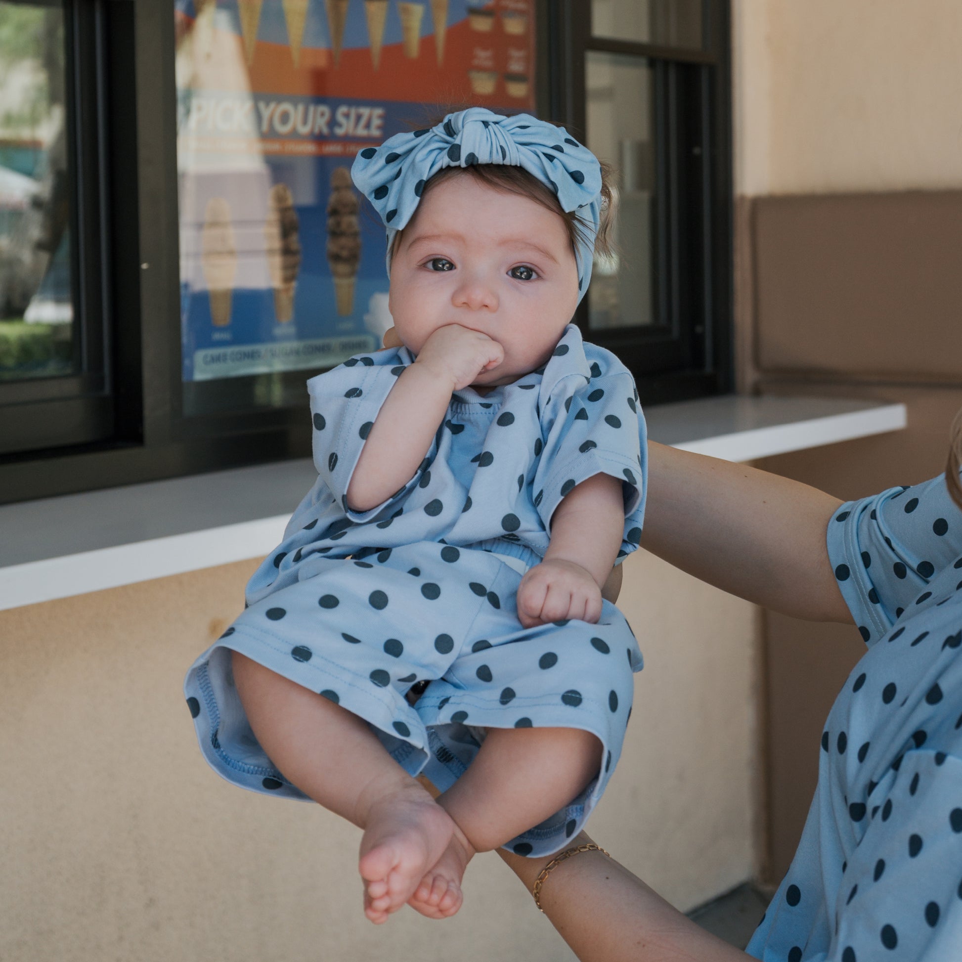 A baby wearing a blue polka-dotted outfit and the Bamboo Head Wrap | Sully from forever french baby is held up by an adult. The baby has a hand in their mouth, adding to the adorable moment. An ice cream poster is visible in the background, perfectly capturing this sweet forever french baby scene.