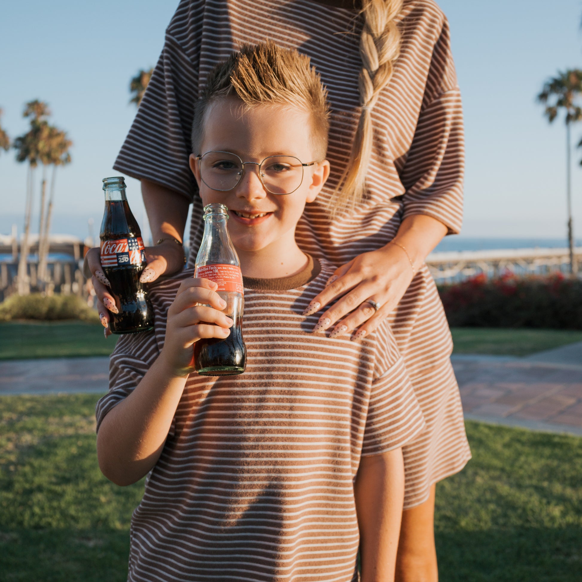 A smiling boy wears the forever french baby Child Terry Short Set | Cola Stripe, holding a Coke bottle beside a woman in a matching dress. They stand outdoors with palm trees, showing off their coordinated family look.