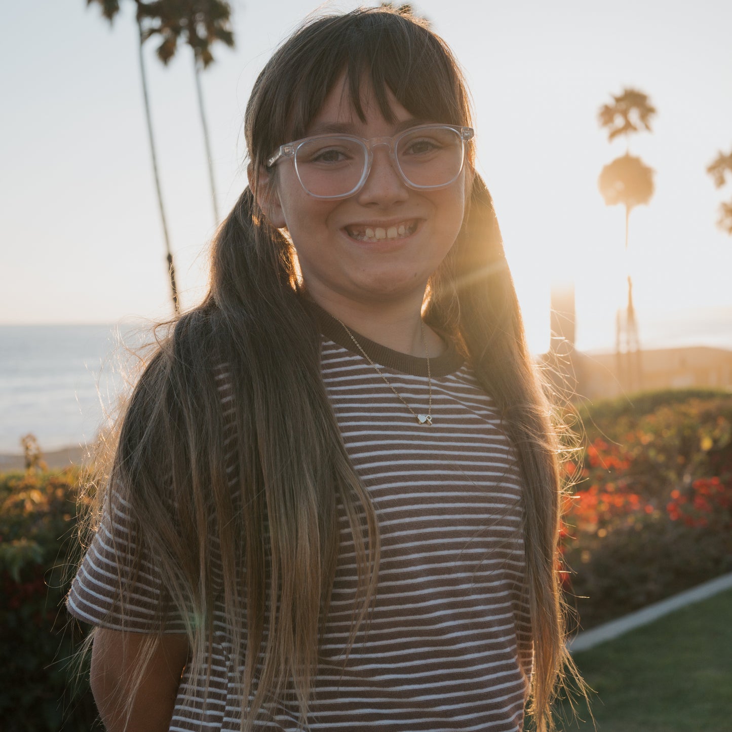 A smiling girl with long brown hair in pigtails, wearing glasses and a striped shirt, stands outdoors at sunset in the forever french baby Child Terry Short Set | Cola Stripe. Palm trees and the ocean are behind her.