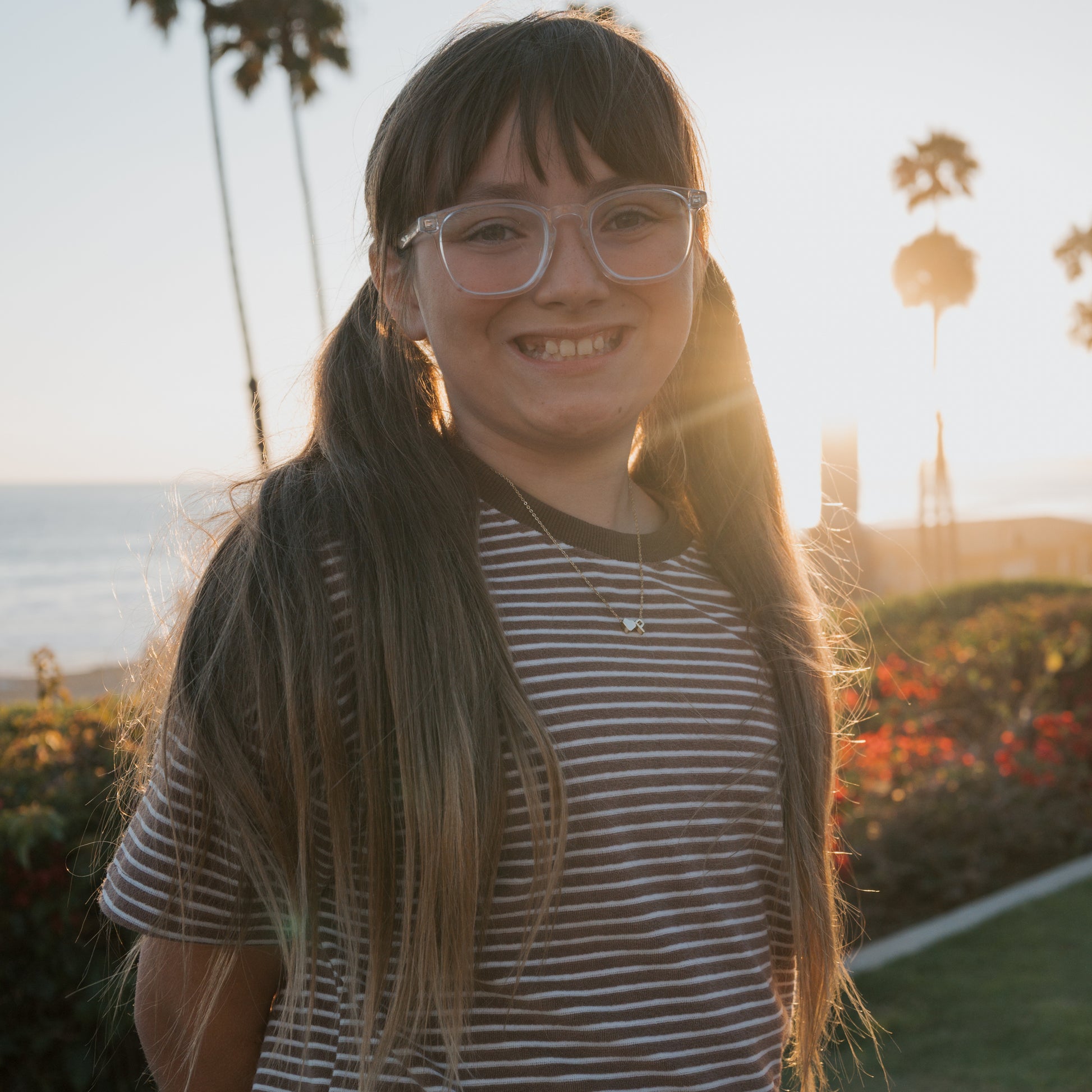 A smiling girl with long brown hair in pigtails, wearing glasses and a striped shirt, stands outdoors at sunset in the forever french baby Child Terry Short Set | Cola Stripe. Palm trees and the ocean are behind her.