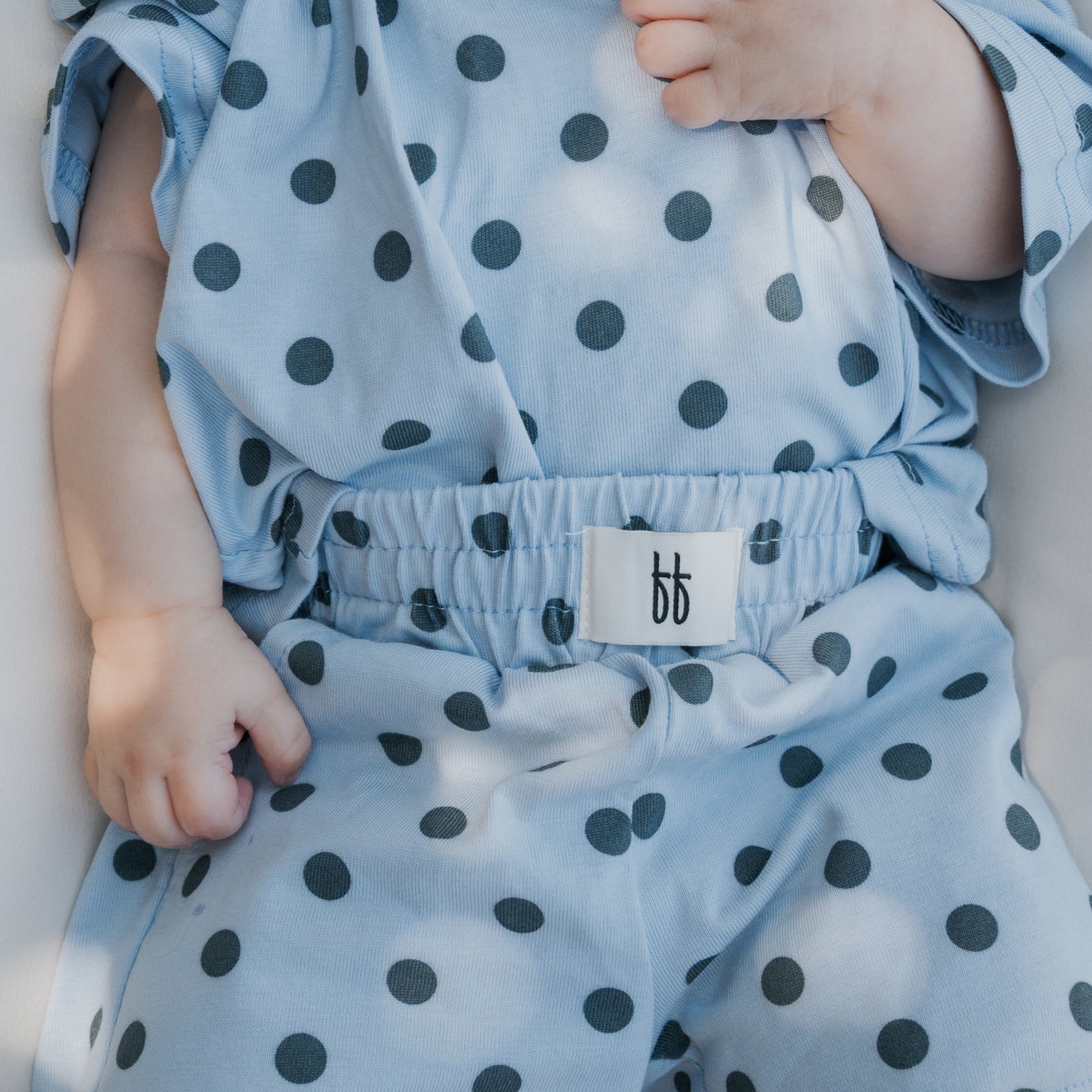 A baby wearing the forever french baby Child Short Set | Sully in light blue cotton with black polka dots, gently holding the fabric near their chest. The set features an elastic waistband and a white tag displaying two lowercase “b” letters—perfect for family matching outfits or as a charming addition to your collection of baby rompers.