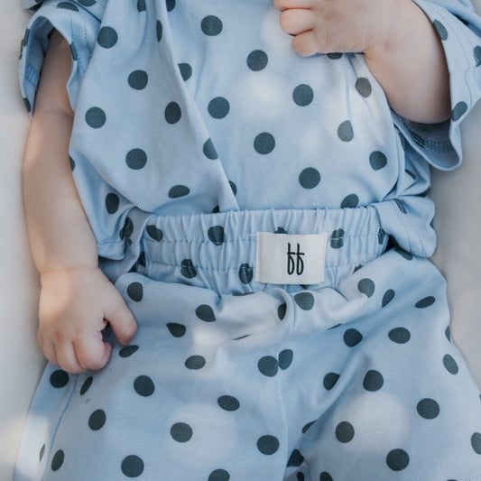 A baby wearing the forever french baby Child Short Set | Sully in light blue cotton with black polka dots, gently holding the fabric near their chest. The set features an elastic waistband and a white tag displaying two lowercase “b” letters—perfect for family matching outfits or as a charming addition to your collection of baby rompers.