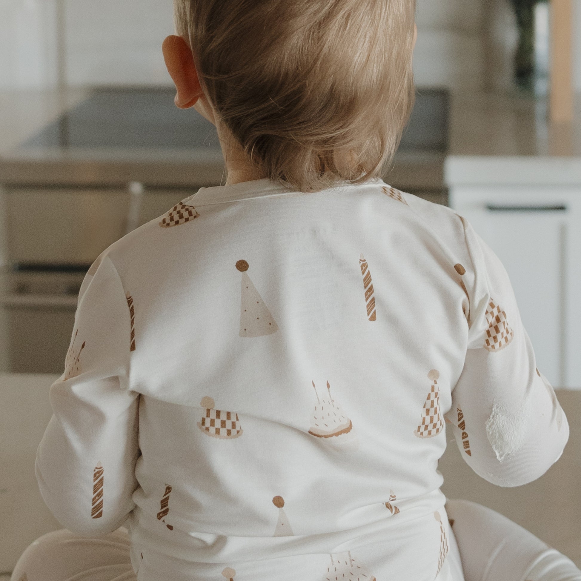 A young child with light brown hair sits on a counter, seen from behind, wearing the forever french baby Child Sleepy Time Set | Birthday Boy—breathable kids pajamas decorated with party hats, candles, and cakes.
