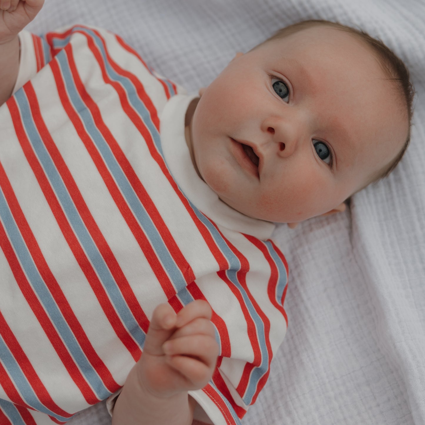 A baby wearing the Child Short Set | Heritage Stripe by forever french baby lies on a white textured blanket, gazing up with wide blue eyes and rosy cheeks.