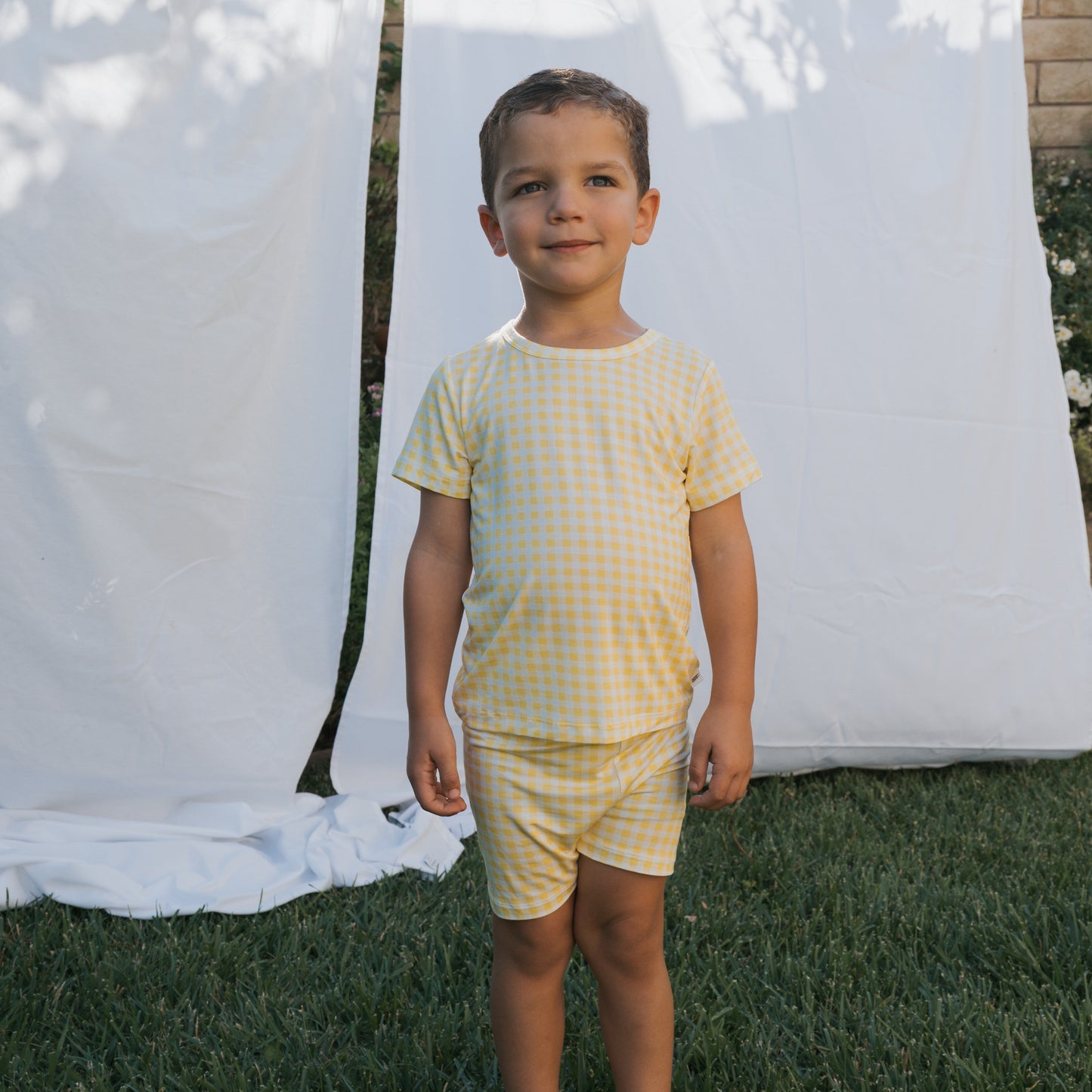 A young boy stands on grass, wearing the Bamboo Short Set | Citron by forever french baby—a breathable yellow and white checkered children's outfit. Behind him, two white sheets hang outside in sunlight, creating a serene backdrop.