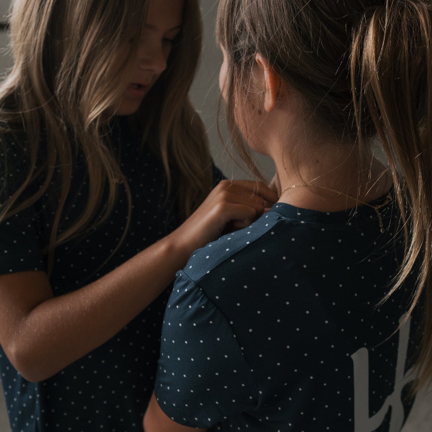 Two girls with long hair, both in forever french baby’s Child Short Sleeve Bamboo Lounge Set in Emery, stand close indoors—one facing the camera and the other gently touching her breathable bamboo clothing.
