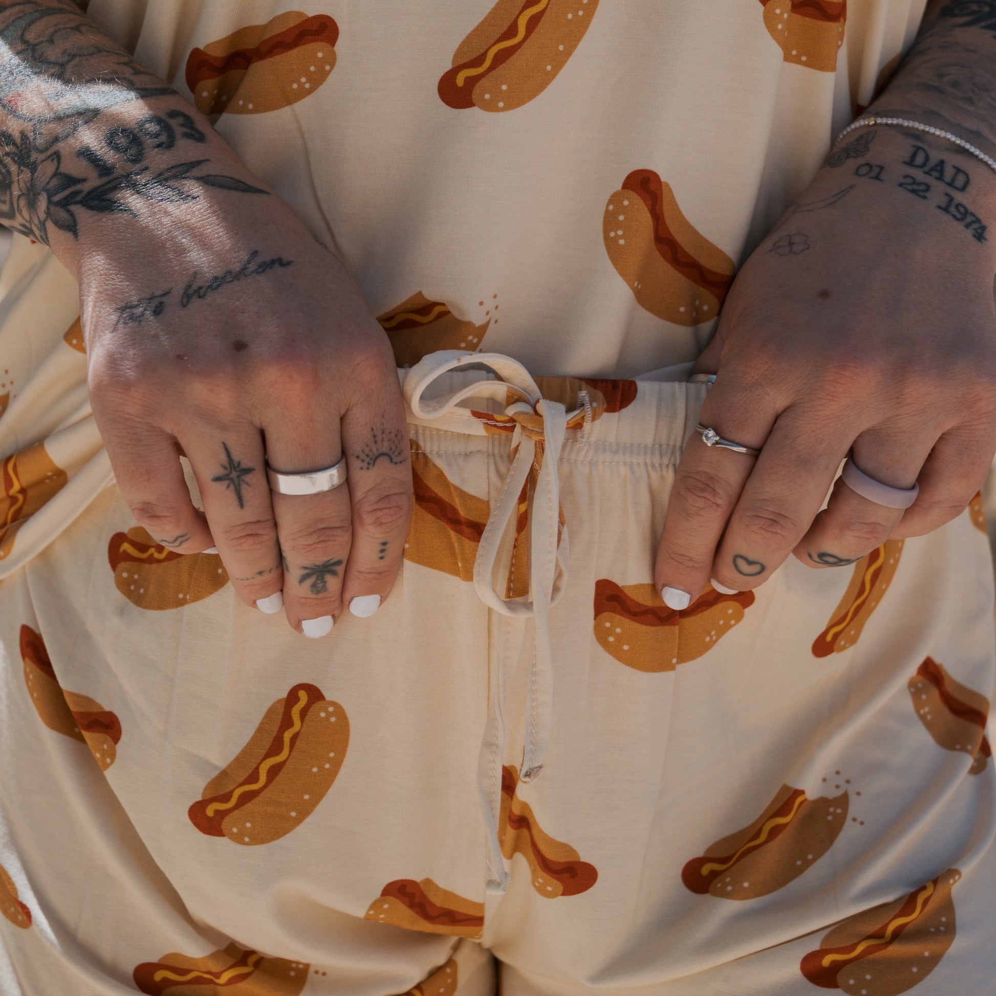 A person with tattooed hands and white nail polish holds the drawstring of forever french baby Women's Bamboo Short Set | Hot Dogs. The beige hot dog print shorts match their shirt; only their torso and hands are visible in the cropped image.