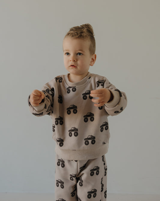 A young child with light brown hair stands against a plain background, wearing the forever french baby Child Sweat Set | Monster Truck, a beige outfit featuring a fun monster truck print—perfect for everyday adventures.