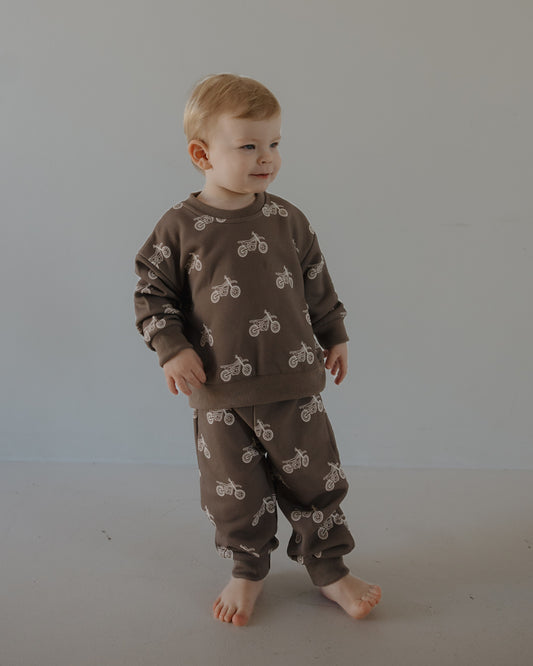 A young child stands barefoot on a gray floor, wearing the forever french baby Child Sweat Set | Dirt Bike in brown with playful white bicycle prints, set against a plain light gray background.