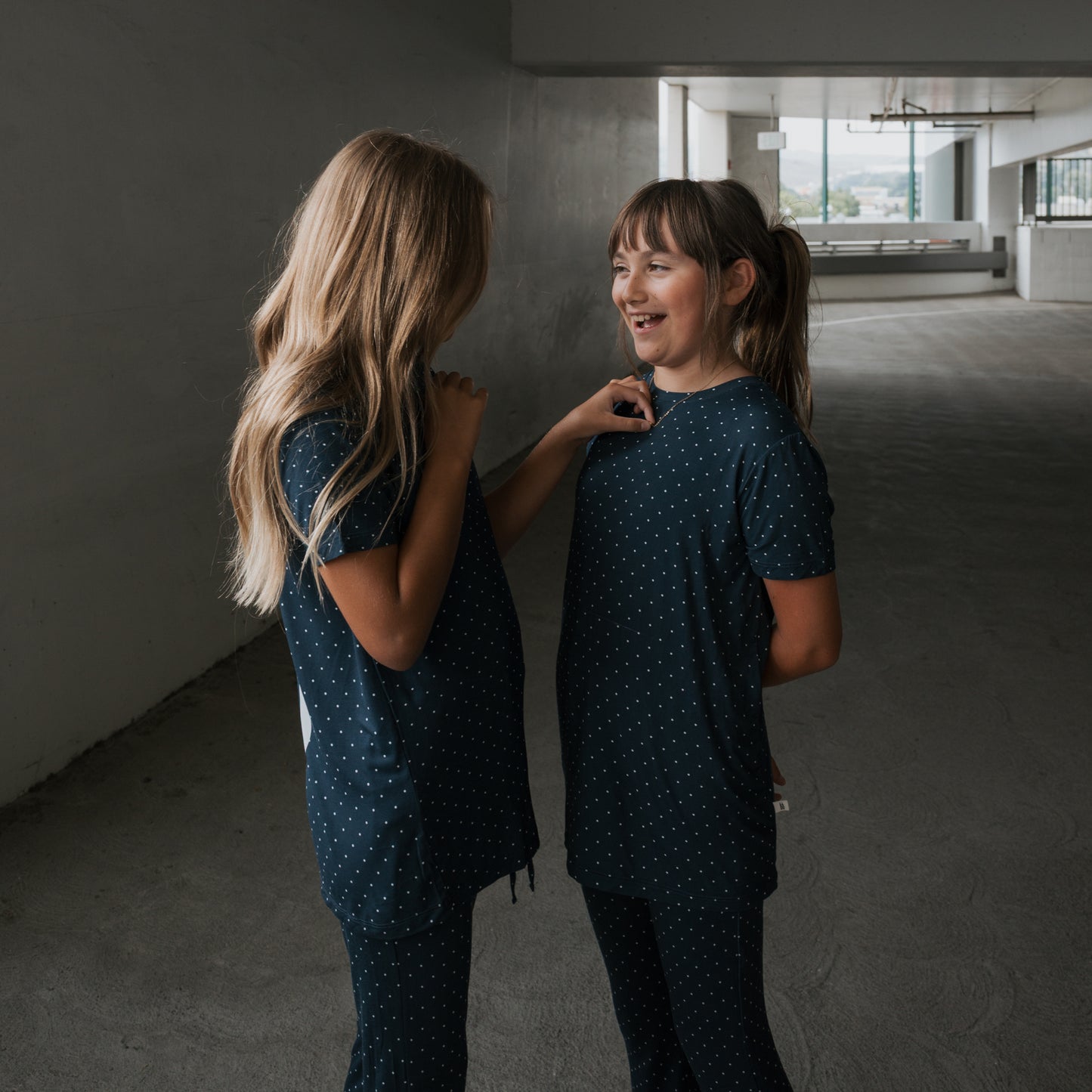 Two girls in matching forever french baby Child Short Sleeve Bamboo Lounge Set | Emery outfits face each other indoors, smiling and enjoying a conversation in their breathable kids’ clothing.