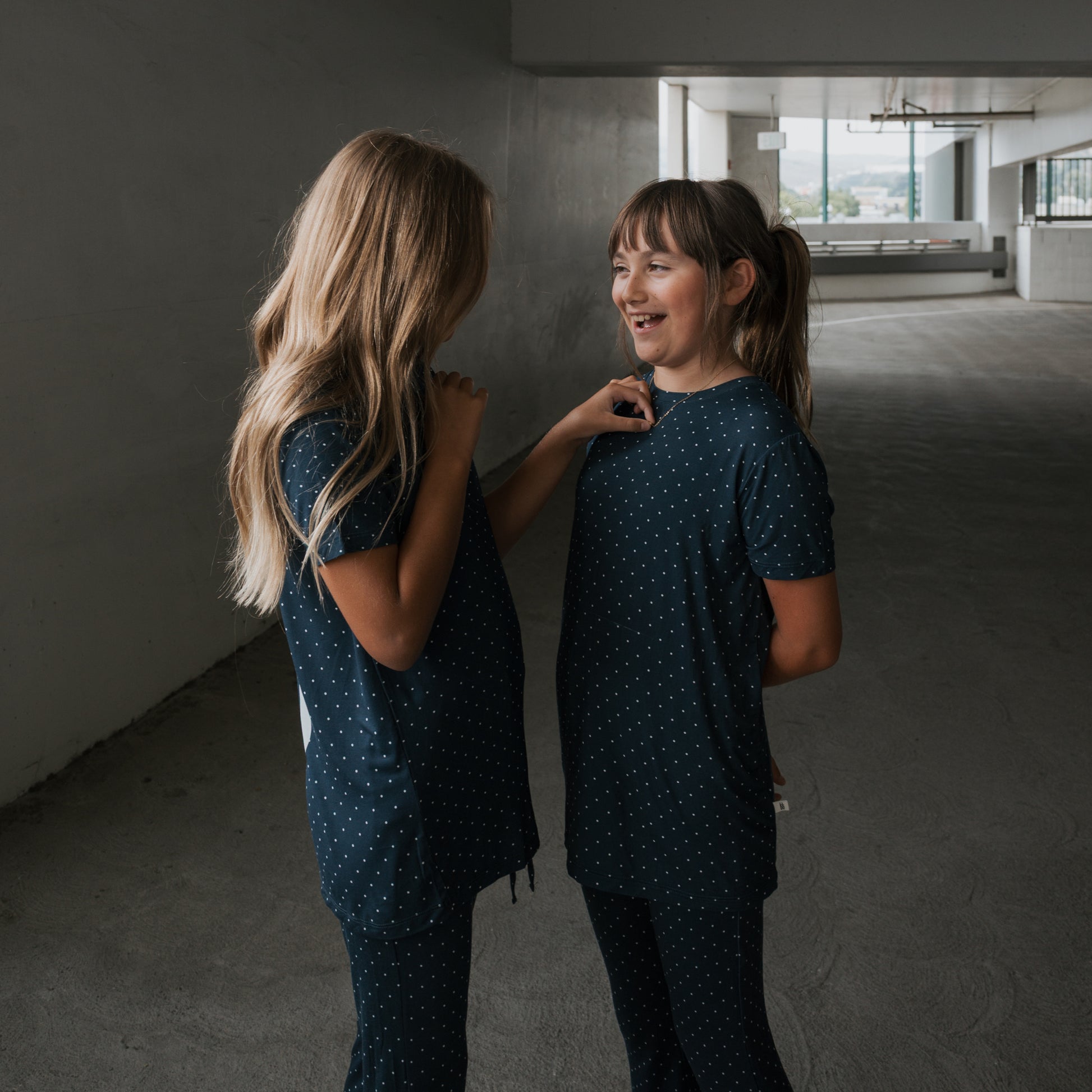 Two girls in matching forever french baby Child Short Sleeve Bamboo Lounge Set | Emery outfits face each other indoors, smiling and enjoying a conversation in their breathable kids’ clothing.