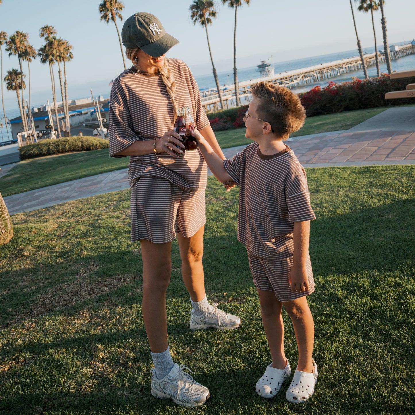 A woman and young boy wear matching outfits by forever french baby near the ocean. The boy wears the Child Terry Short Set in Cola Stripe. Palm trees, a pier, and a bench are in the background as they smile at each other.