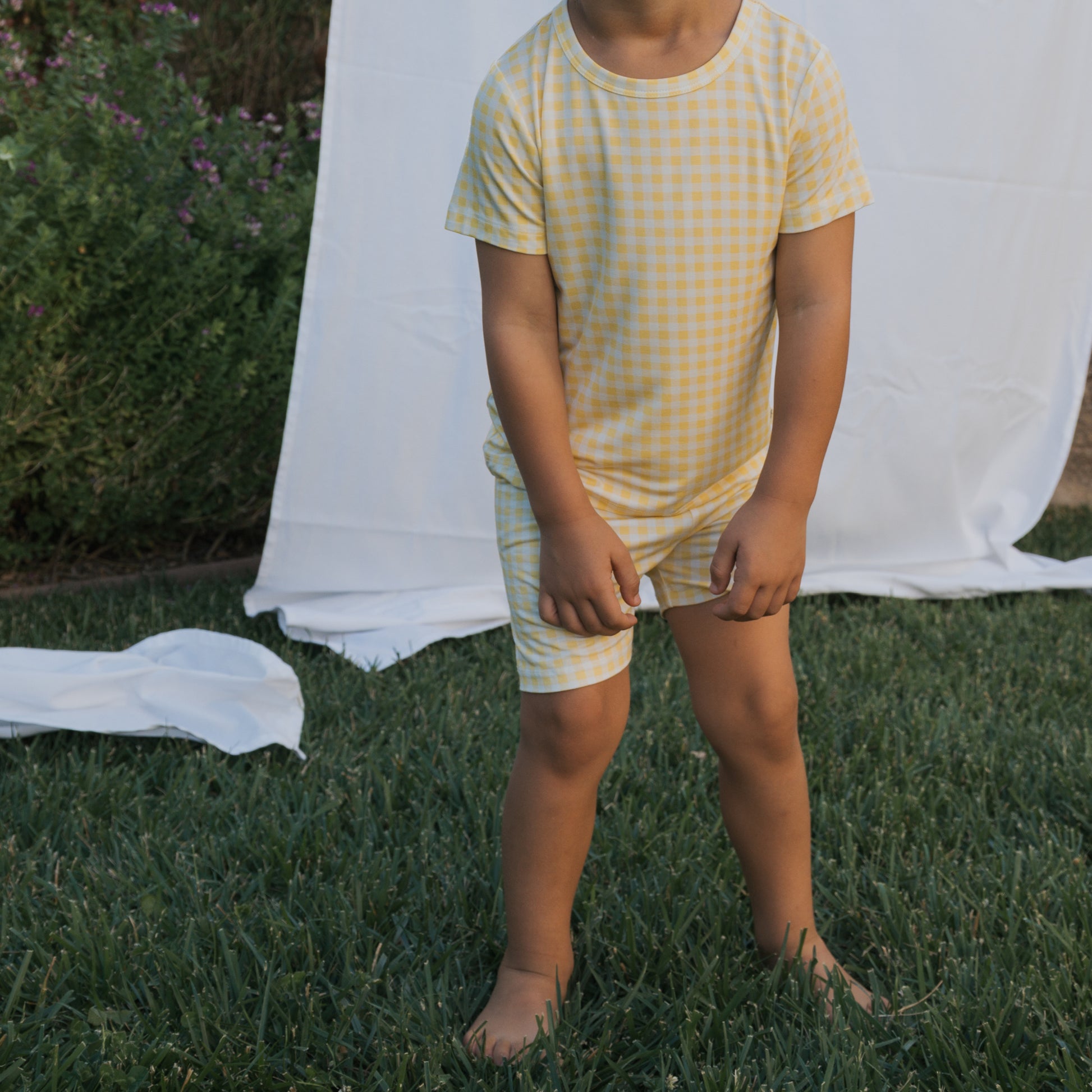 A child stands on grass before a white sheet backdrop, wearing the forever french baby Bamboo Short Set | Citron. The yellow and white checkered set is breathable; the face is hidden. Green plants with purple flowers appear in the background.