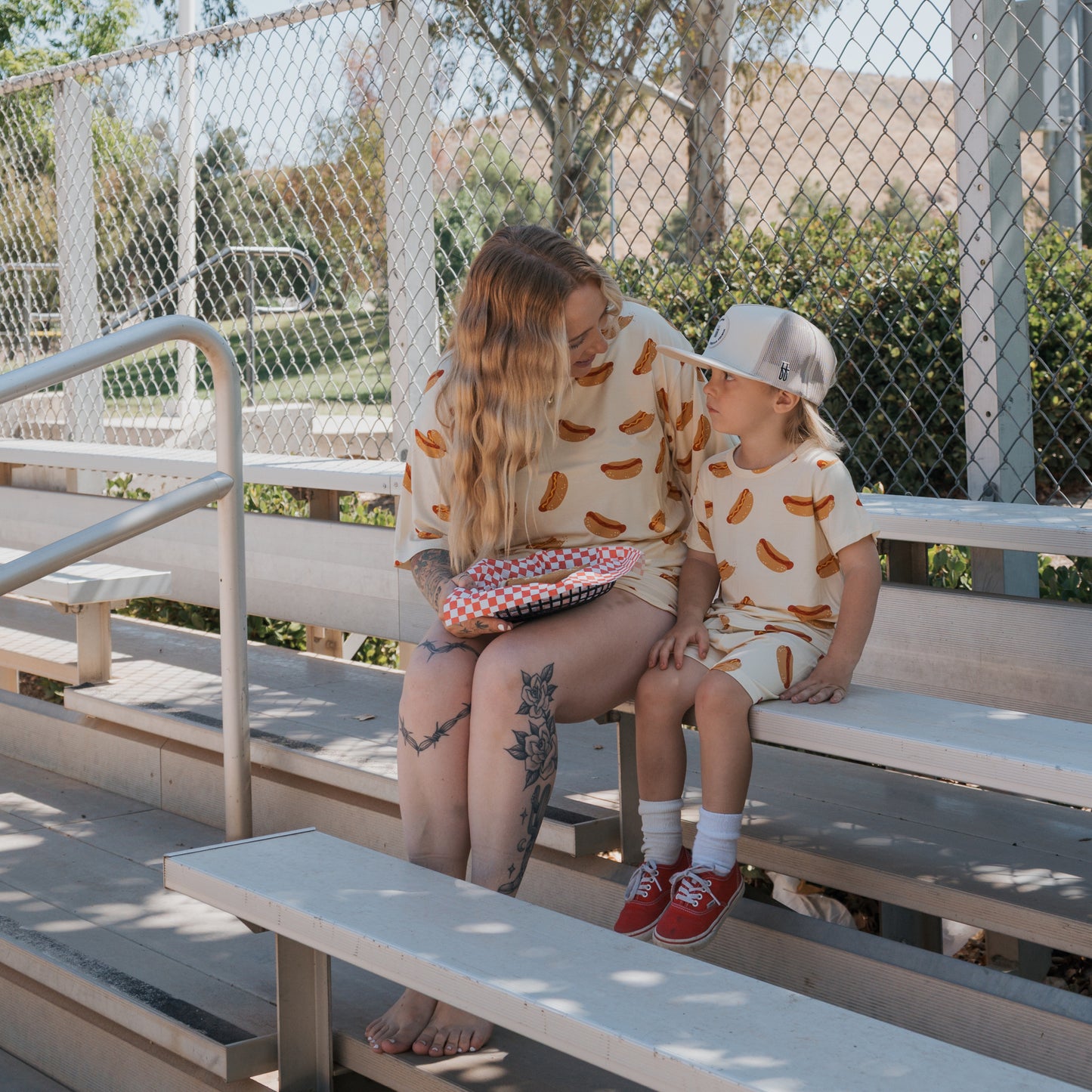 A woman and young child wearing matching forever french baby Bamboo Short Sets in the Hot Dogs print smile at each other on outdoor bleachers. The woman has long hair and tattoos, while the child wears a cap and red shoes.