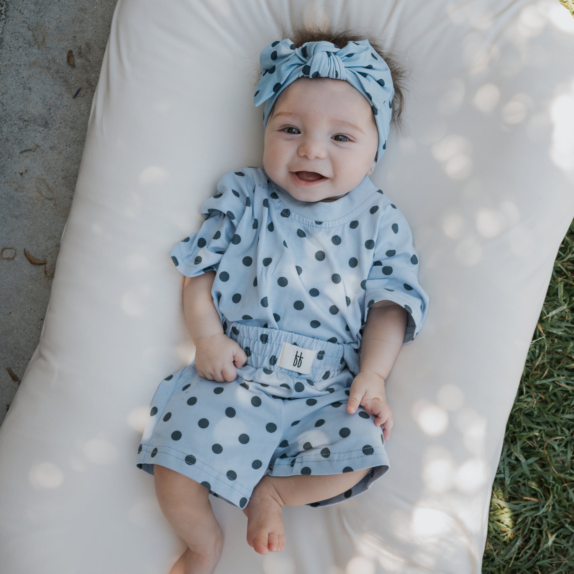 A baby wearing the forever french baby Child Short Set | Sully—a light blue polka dot romper and matching headband—lies on a white cushion outdoors, smiling with hands and feet visible in dappled sunlight. Ideal for family matching outfits or as a comfy cotton child short set.