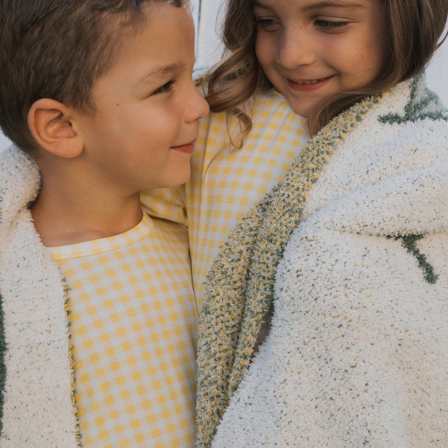 Two children wearing forever french baby’s Bamboo Short Set in Citron smile at each other, wrapped in a white and green blanket. Their warm expressions reflect the comfort of breathable bamboo children’s clothing.