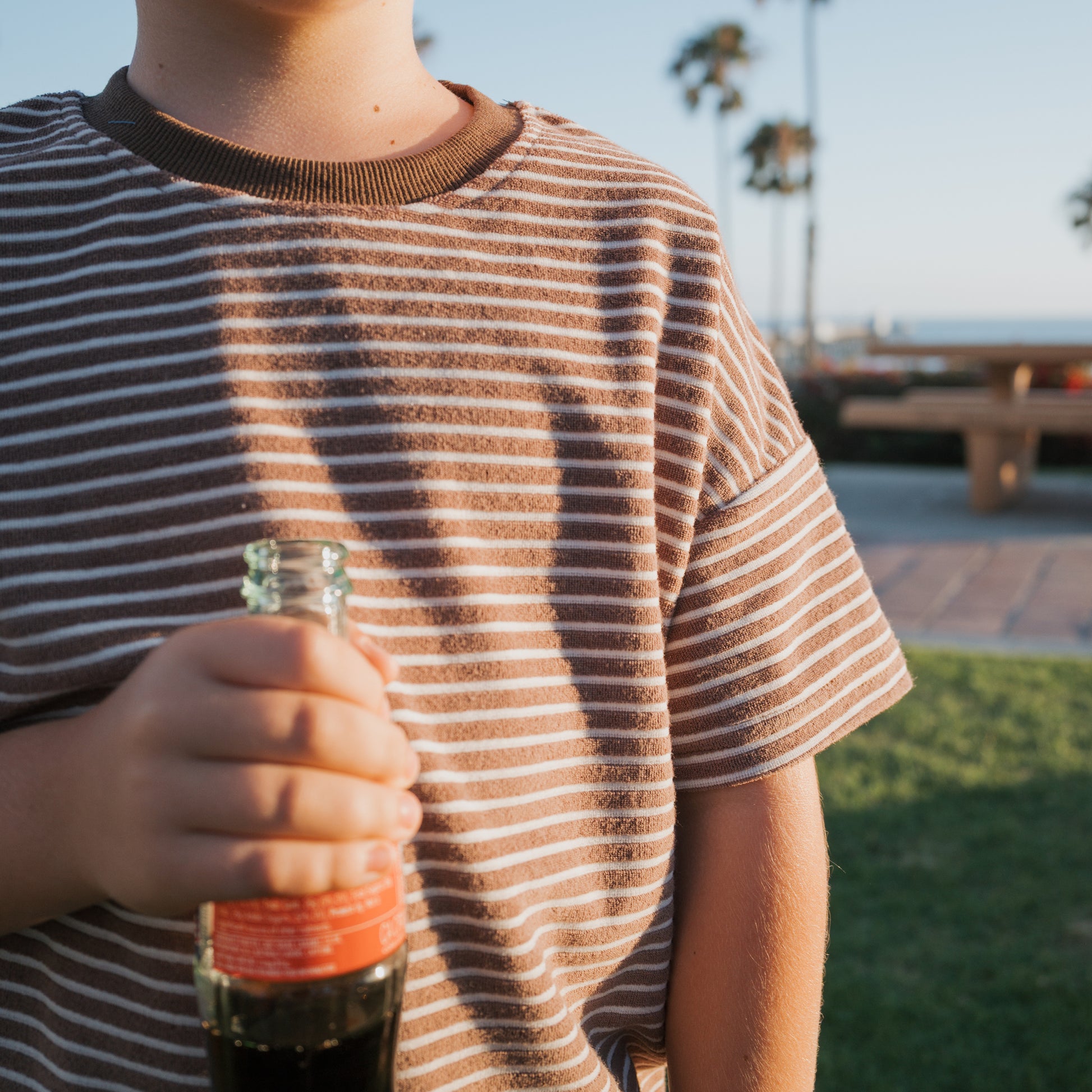 A child wears the forever french baby Child Terry Short Set in Cola Stripe and holds a glass soda bottle outdoors, with palm trees, a bench, and blue sky suggesting family matching outfits for a sunny park or beach day.