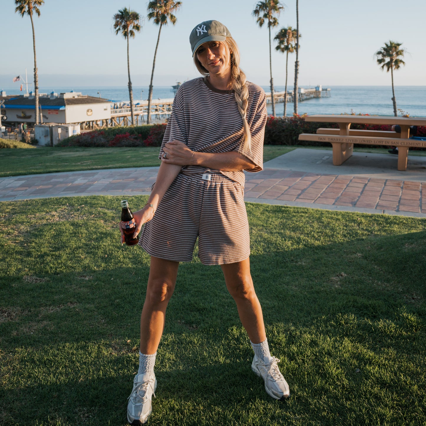 Wearing the forever french baby Women's Terry Short Set in Cola Stripe, a person stands on grass with a glass bottle; sneakers and a baseball cap complete the look. Palm trees, a wooden picnic table, and a beach pier appear in the background.