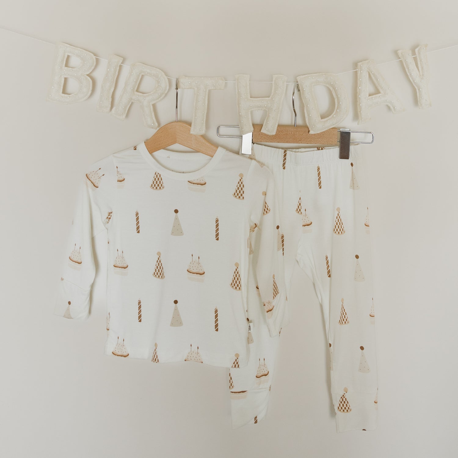 The Child Sleepy Time Set | Birthday Boy by forever french baby, a two-piece breathable kids pajama set featuring birthday-themed prints, hangs on wooden hangers. Behind the pajamas, white soft letters spell “BIRTHDAY” on the wall as a festive decoration, creating a cheerful scene perfect for celebrating in comfort.
