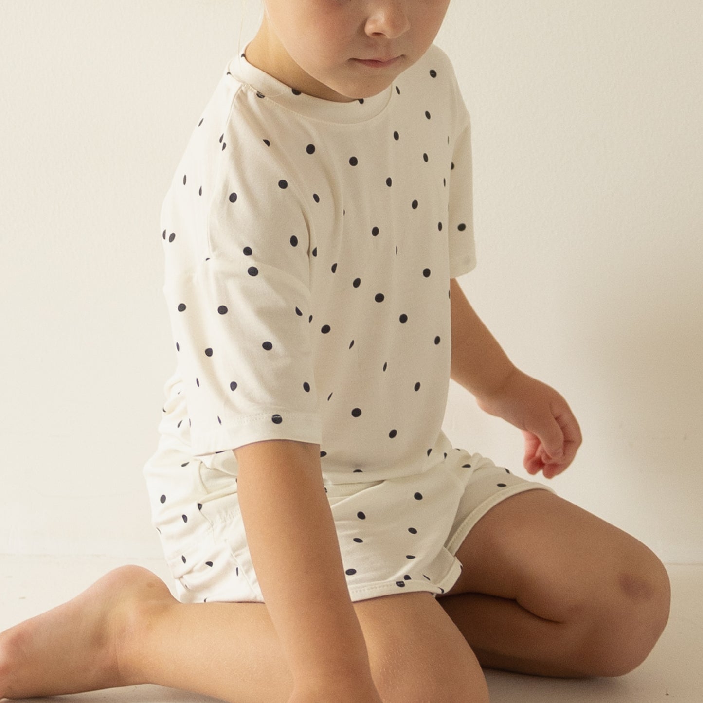 A child wears the forever french baby Girl's Bamboo Short Set | Annie—a white polka dot shirt and shorts—sitting on the floor against a plain white background, looking slightly downward.