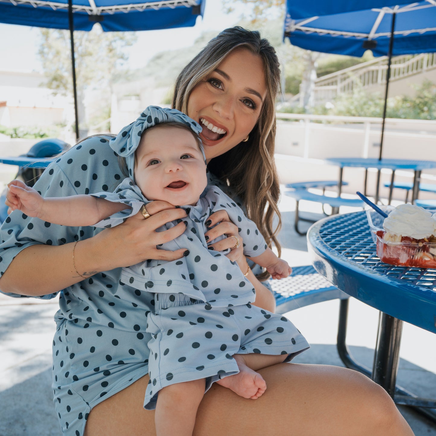 A smiling woman holds a happy baby; both are dressed in matching blue polka-dot outfits from forever french baby. Her child is wearing the adorable "Child Short Set | Sully" while they sit together at a blue picnic table topped with dessert, under blue umbrellas outdoors, perfectly showcasing their charming family matching outfits.