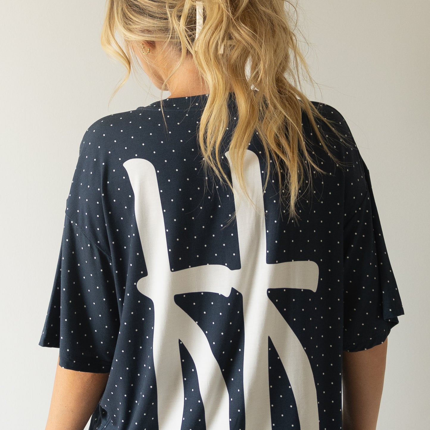 A person with long blonde hair in a ponytail, facing away from the camera, wears the forever french baby Short Sleeve Women's Bamboo Lounge Set | Emery in dark blue with white polka dots and a large abstract white design on the back.