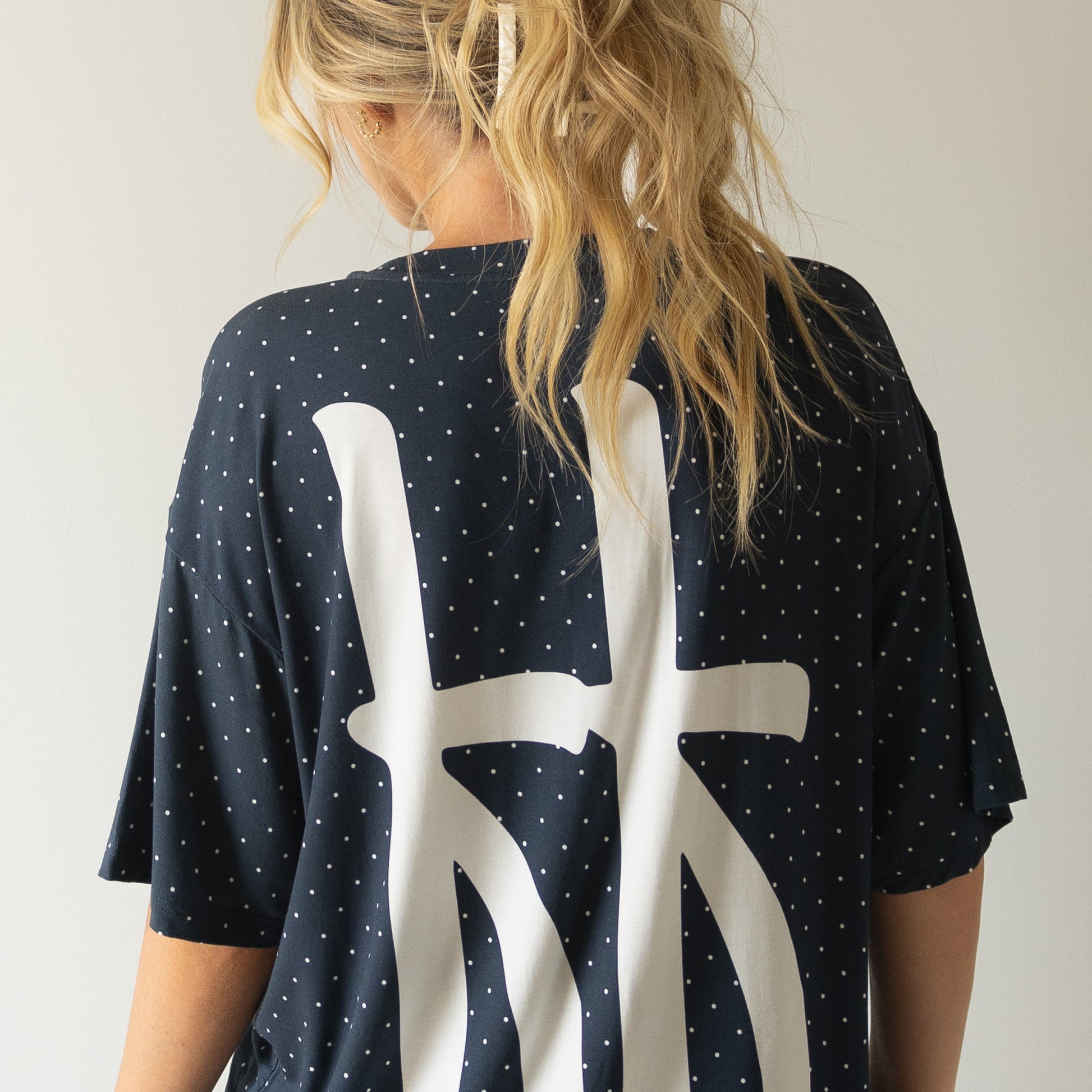 A person with long blonde hair in a ponytail, facing away from the camera, wears the forever french baby Short Sleeve Women's Bamboo Lounge Set | Emery in dark blue with white polka dots and a large abstract white design on the back.