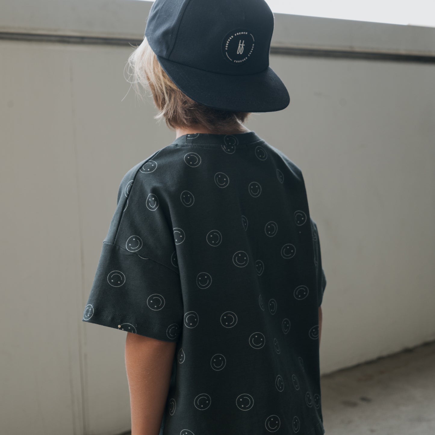 A child wearing the forever french baby Child Short Set | Smiley, featuring a dark top with small smiley faces and a black cap, stands with their back to the camera in a bright indoor space.