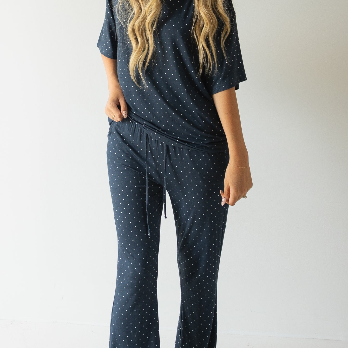 A woman with long wavy blonde hair wears the forever french baby Short Sleeve Women's Bamboo Lounge Set | Emery in navy blue with white polka dots and drawstring pants, standing before a plain light background.