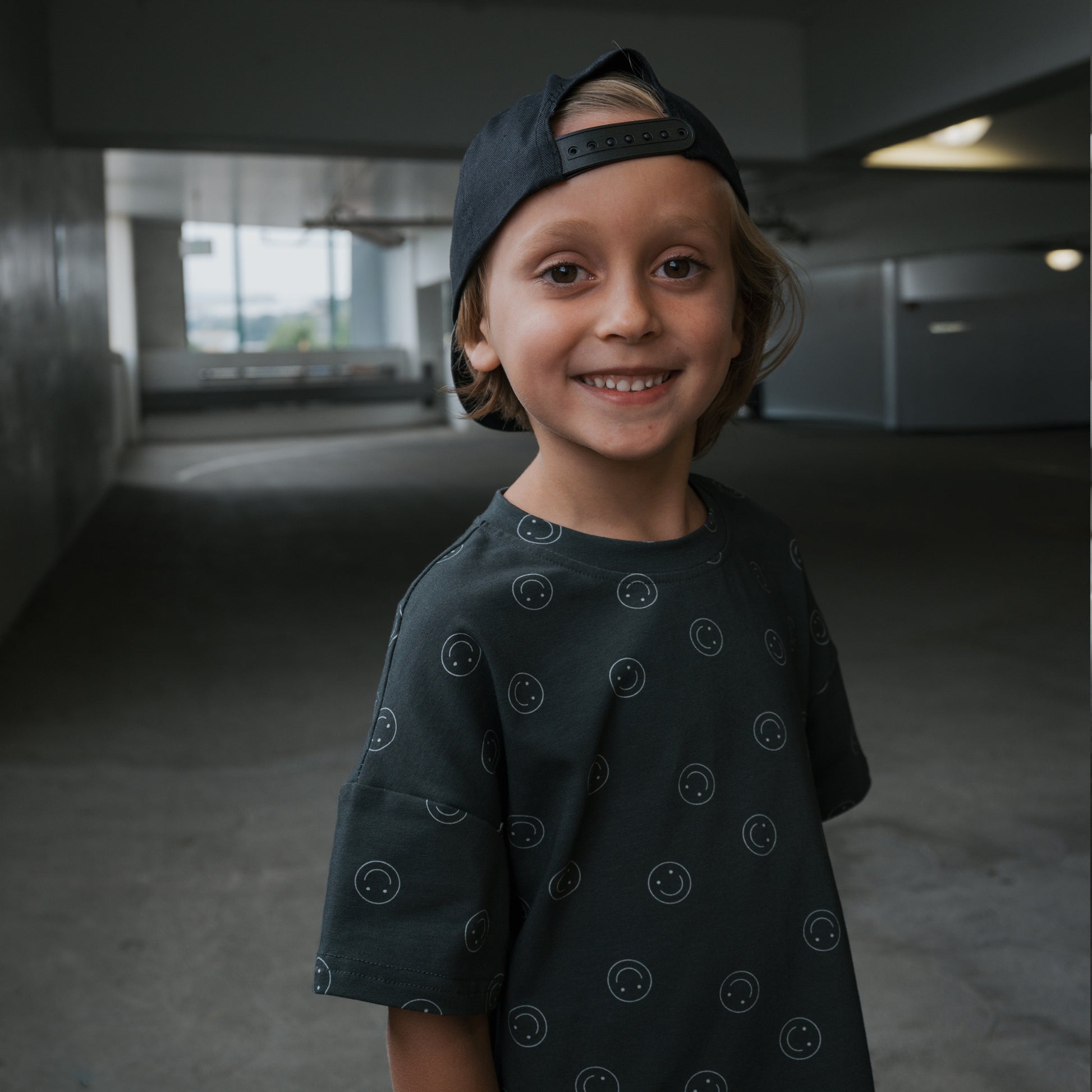 A young boy smiles in a dimly lit parking garage wearing the forever french baby Child Short Set | Smiley, featuring a dark cotton-blend top with smiley face patterns and a backward black cap.
