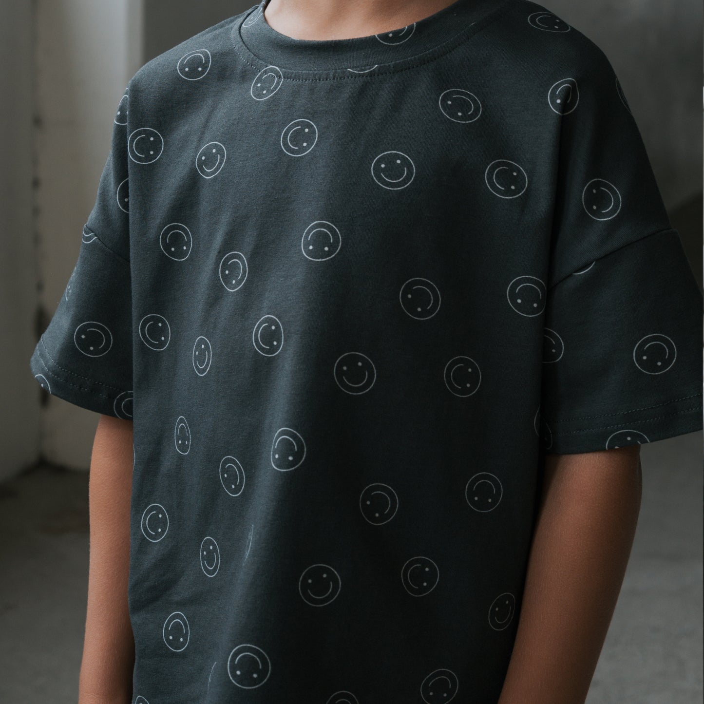 A child models the forever french baby Child Short Set | Smiley, showing a dark gray cotton blend T-shirt with small white smiley faces. The indoor photo is cropped to highlight the upper body, with the head partially out of frame.