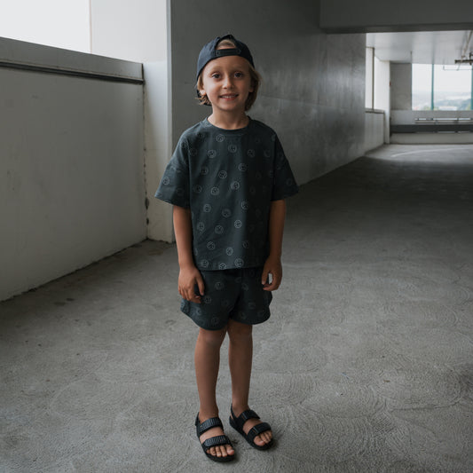 A young child in the forever french baby Child Short Set | Smiley stands smiling in a naturally lit concrete corridor, wearing dark sandals and a backwards cap.