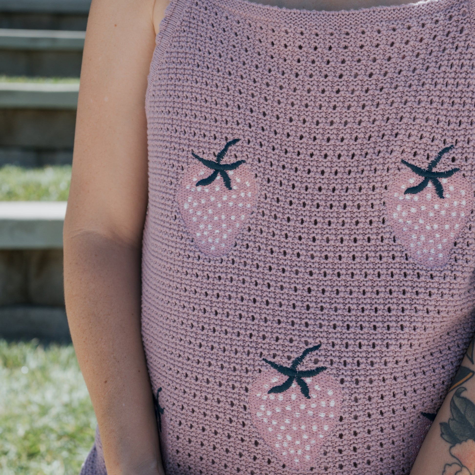 A person wearing the forever french baby Women's Knit Short Set | Matilda—a 100% cotton mauve crochet top adorned with pink strawberries and black stems—stands outdoors near stone steps.