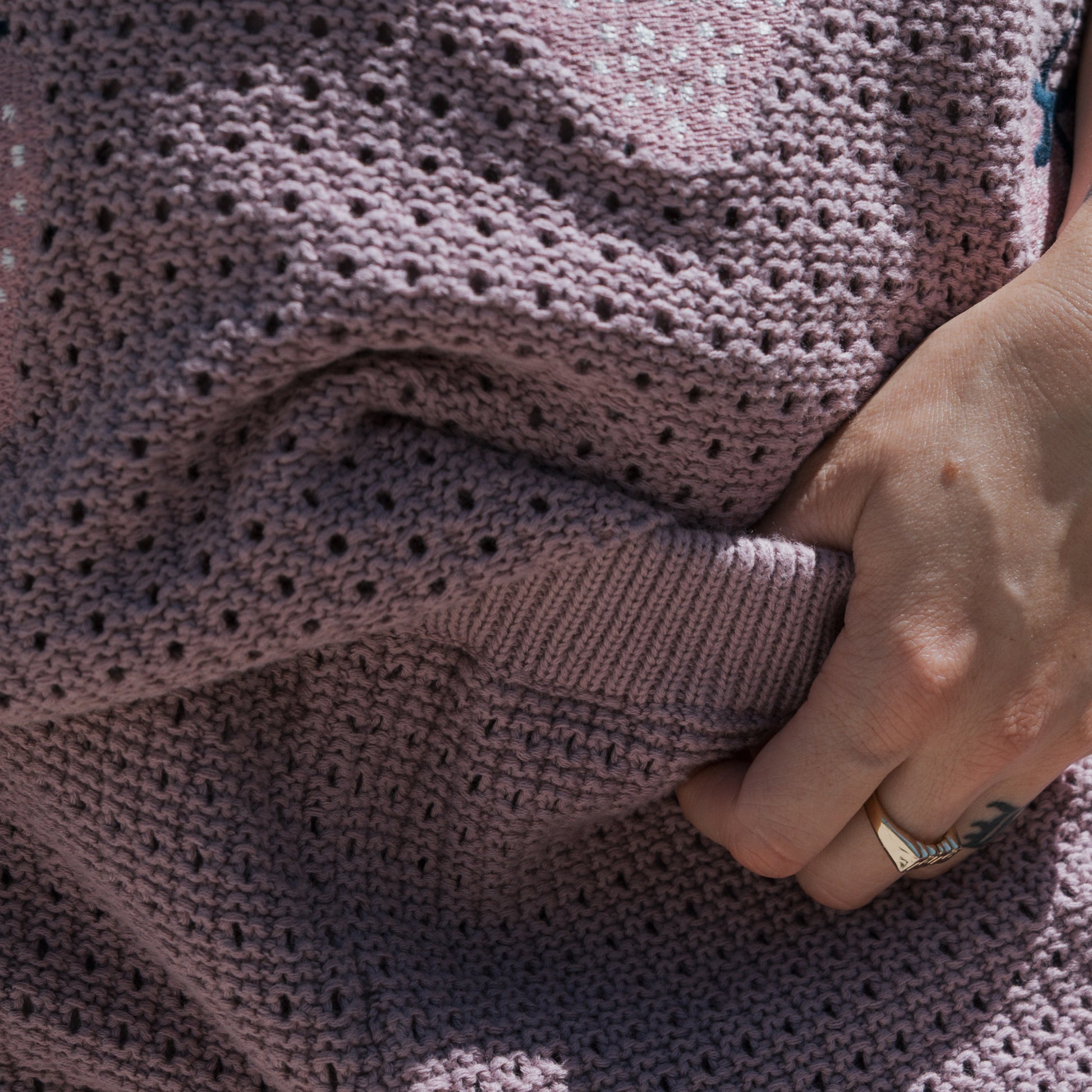 A hand wearing a gold ring gently grasps a textured mauve soft knit fabric, with tattooed fingers partially visible—this cozy 100% cotton piece is the Women's Knit Short Set | Matilda from forever french baby, perfect for pairing with the matching shorts.
