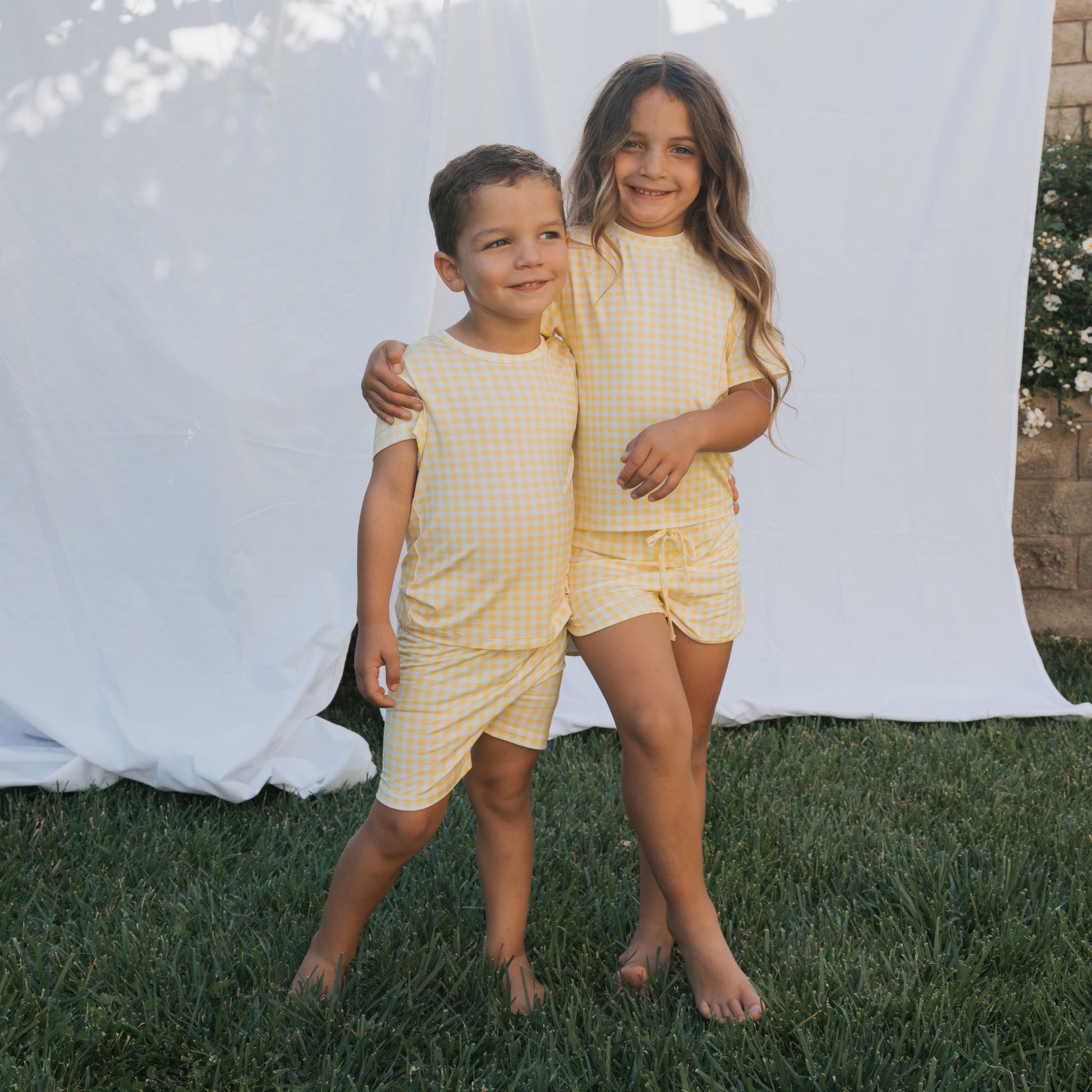 Two young children smile and hug barefoot on grass, wearing matching forever french baby Bamboo Short Set | Citron outfits. The breathable yellow and white gingham clothing stands out against a backdrop of greenery, a brick wall, and a white sheet.