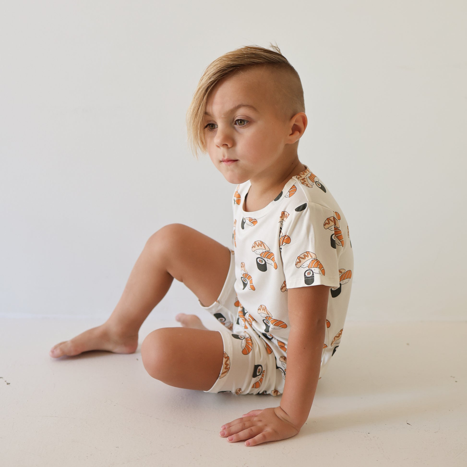A young child with short, side-swept blonde hair sits on the floor in the forever french baby Bamboo Short Set | Sushi—breathable bamboo T-shirt and shorts with sushi prints—against a plain white background.