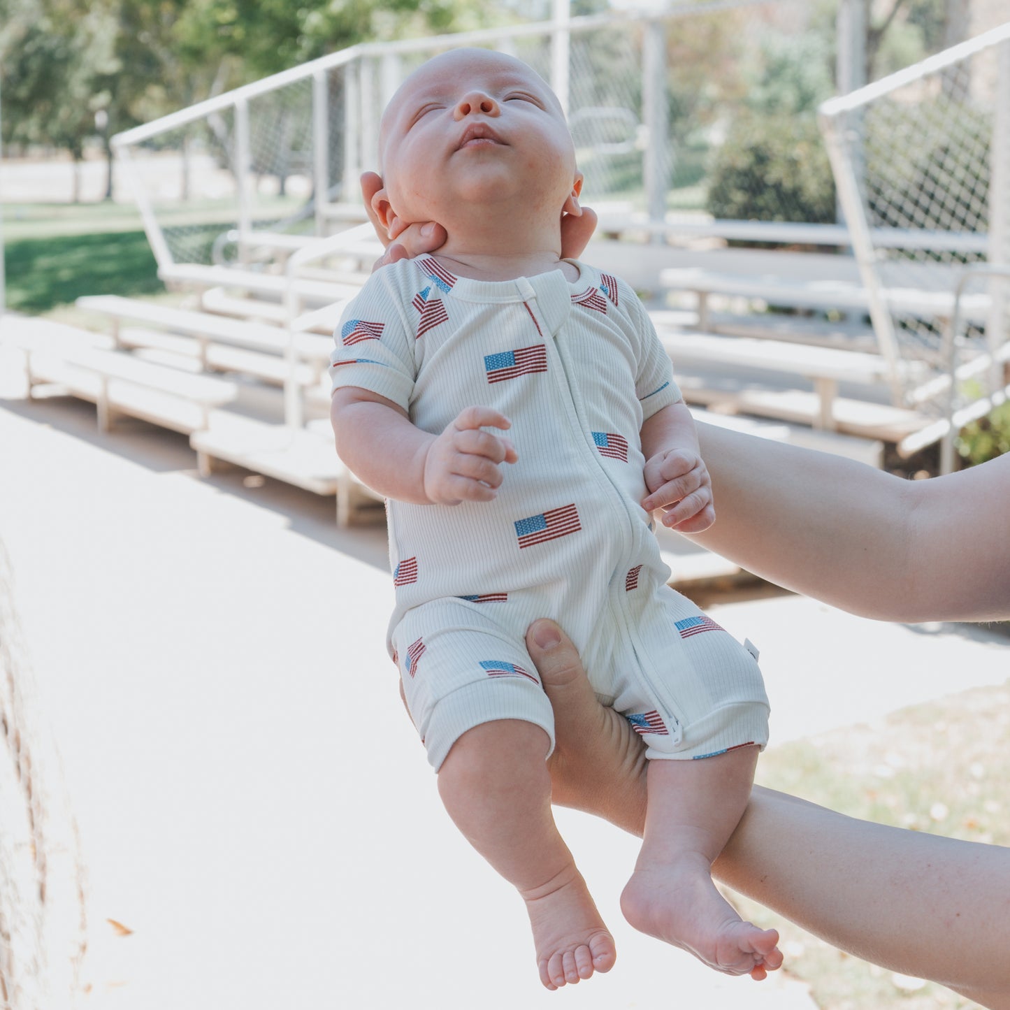 A person holds a baby outdoors, dressed in the forever french baby Shortie Bamboo Zip | Americana Ribbed romper featuring American flags, eyes closed and facing up, with a sunny park and bleachers behind.