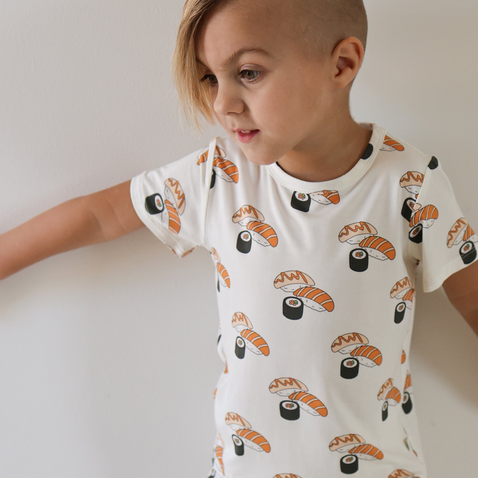 A young child with short, side-shaved blonde hair wears the forever french baby Bamboo Short Set | Sushi, featuring sushi and nigiri prints on a white tee, standing against a plain white wall in soft, breathable bamboo fabric.