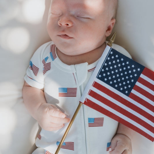 A baby in the forever french baby Shortie Bamboo Zip | Americana Ribbed romper holds a small U.S. flag while lying on a light surface.