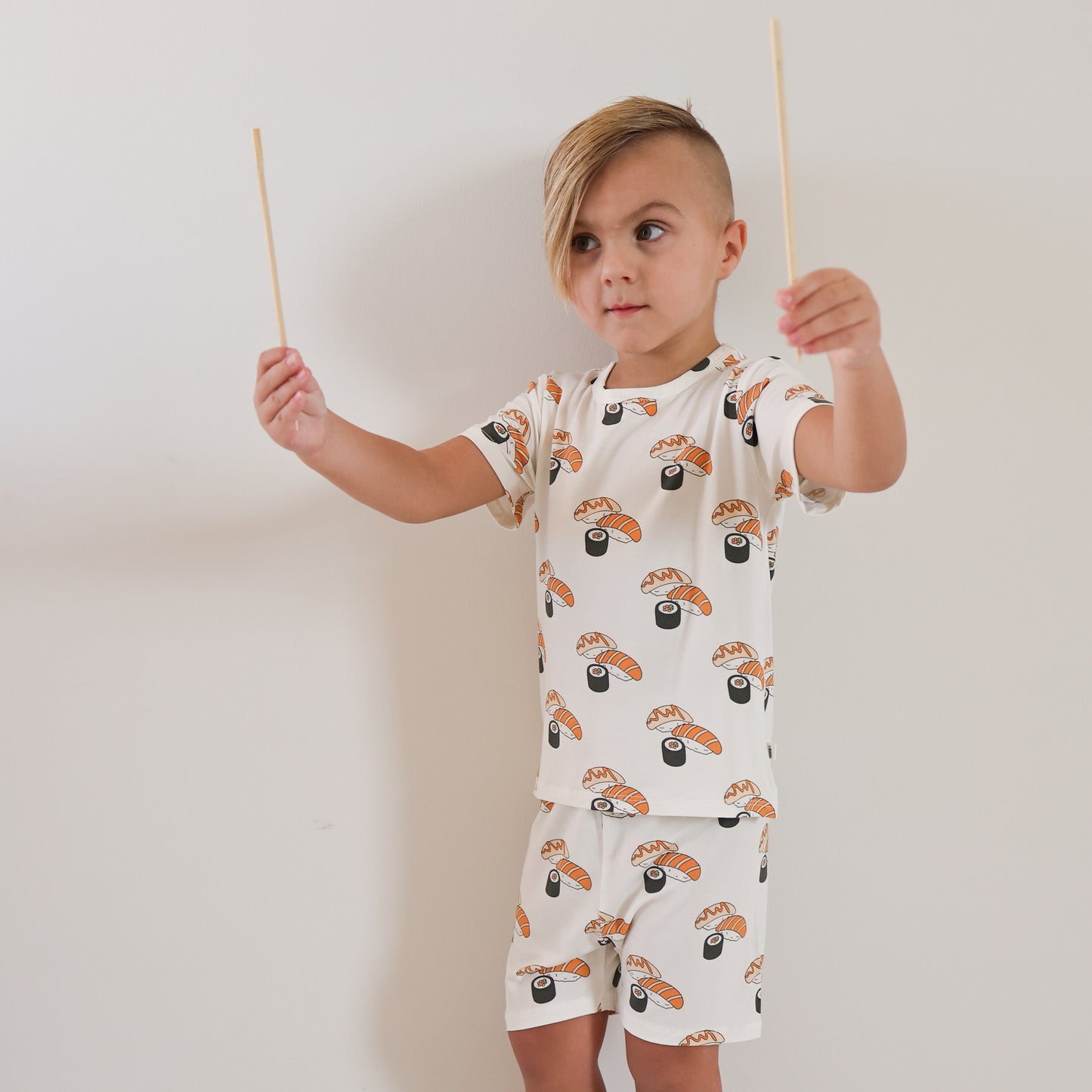 A young child with short, side-swept hair wears the Bamboo Short Set | Sushi by forever french baby and holds two chopsticks in both hands against a plain white wall.
