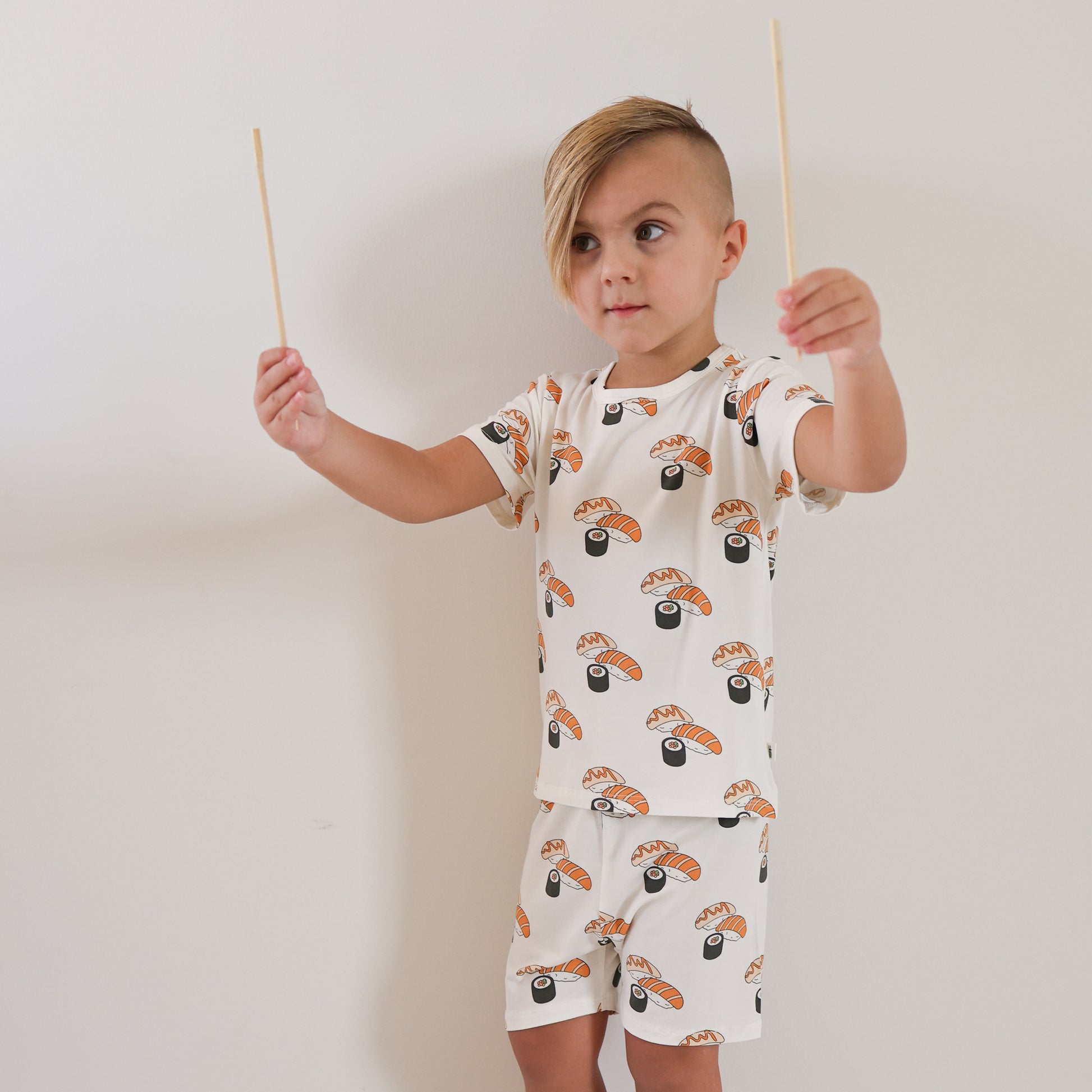 A young child with short, side-swept hair wears the Bamboo Short Set | Sushi by forever french baby and holds two chopsticks in both hands against a plain white wall.