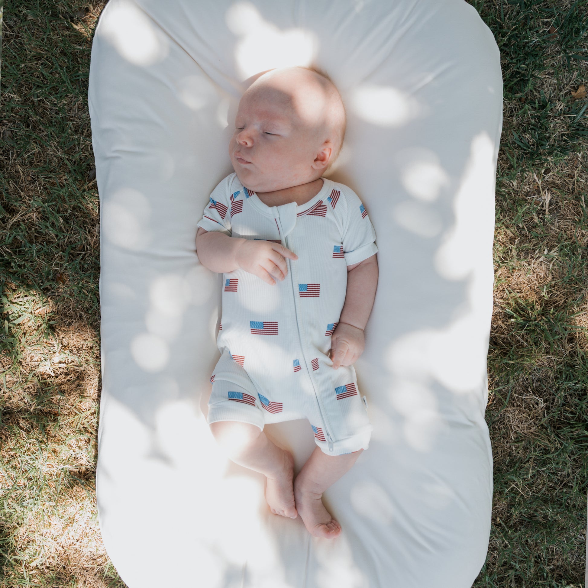 A baby wearing the forever french baby Shortie Bamboo Zip | Americana Ribbed romper sleeps peacefully on a white cushion outdoors, sunlight and tree shadows dancing softly on the grass around them.